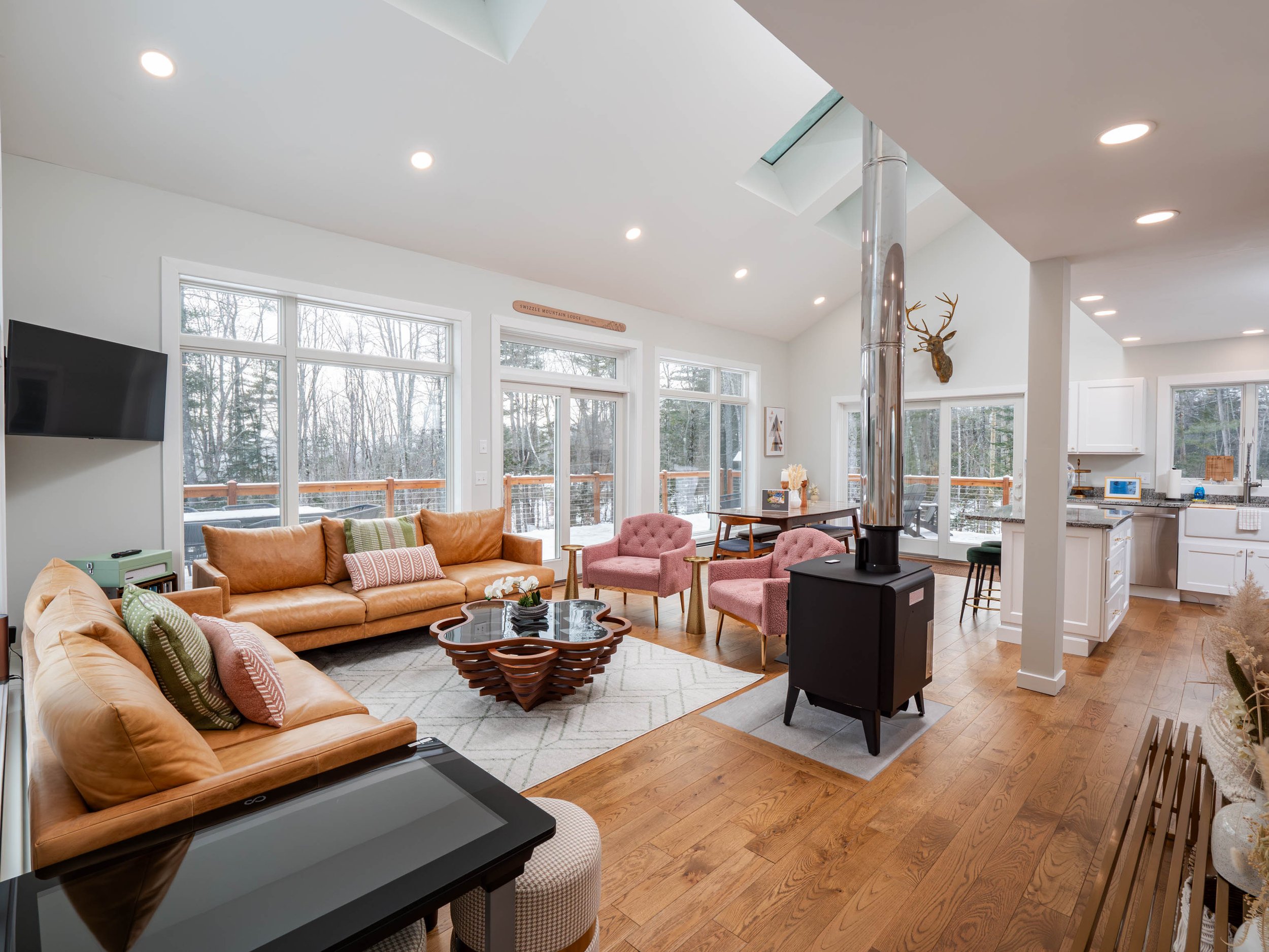 Open-concept living room with large windows, beige and pink sofas, a black stove, wooden flooring, and kitchen in the background.