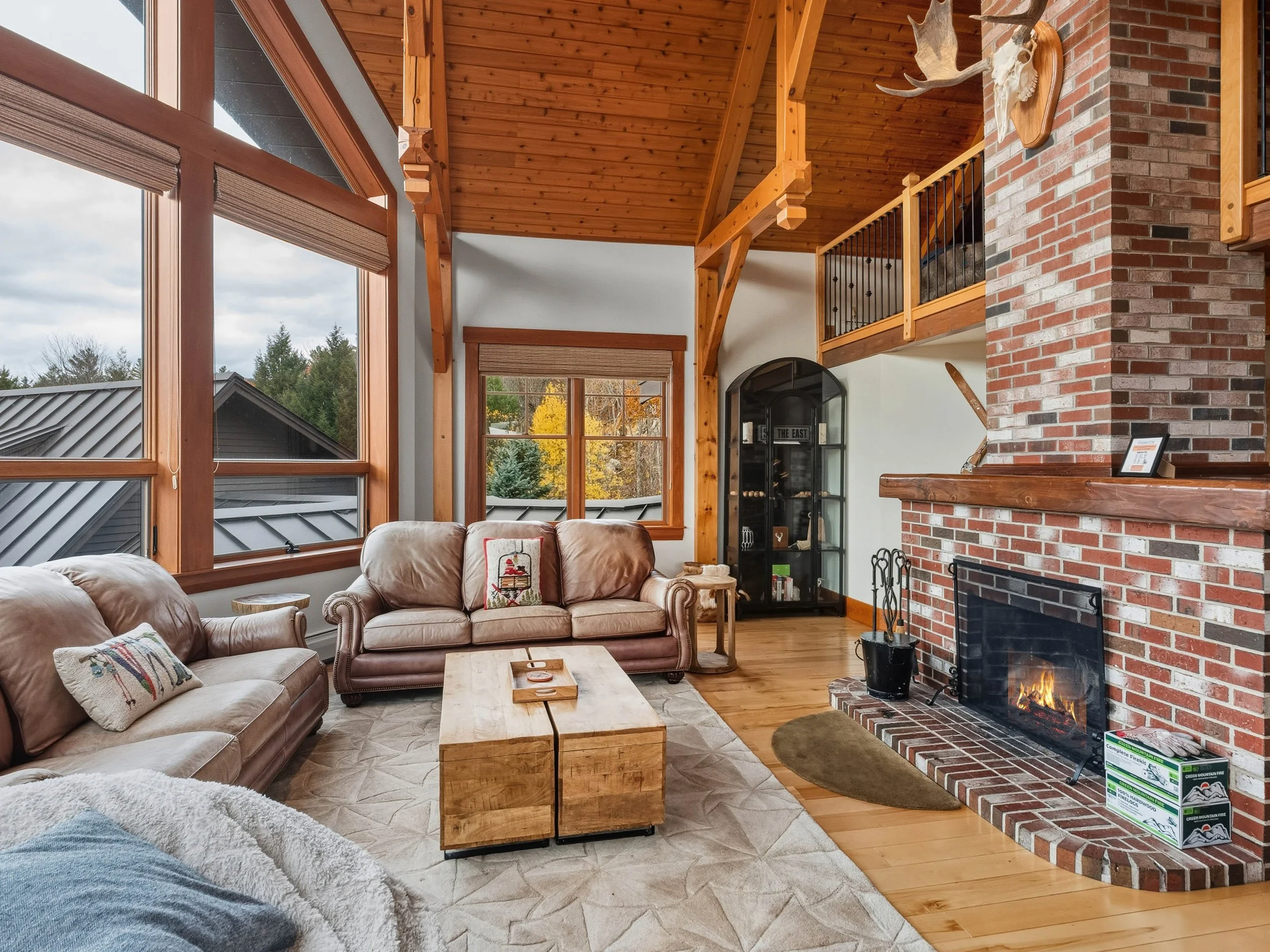 Cozy living room with leather sofas, large windows, a brick fireplace, and wooden accents.