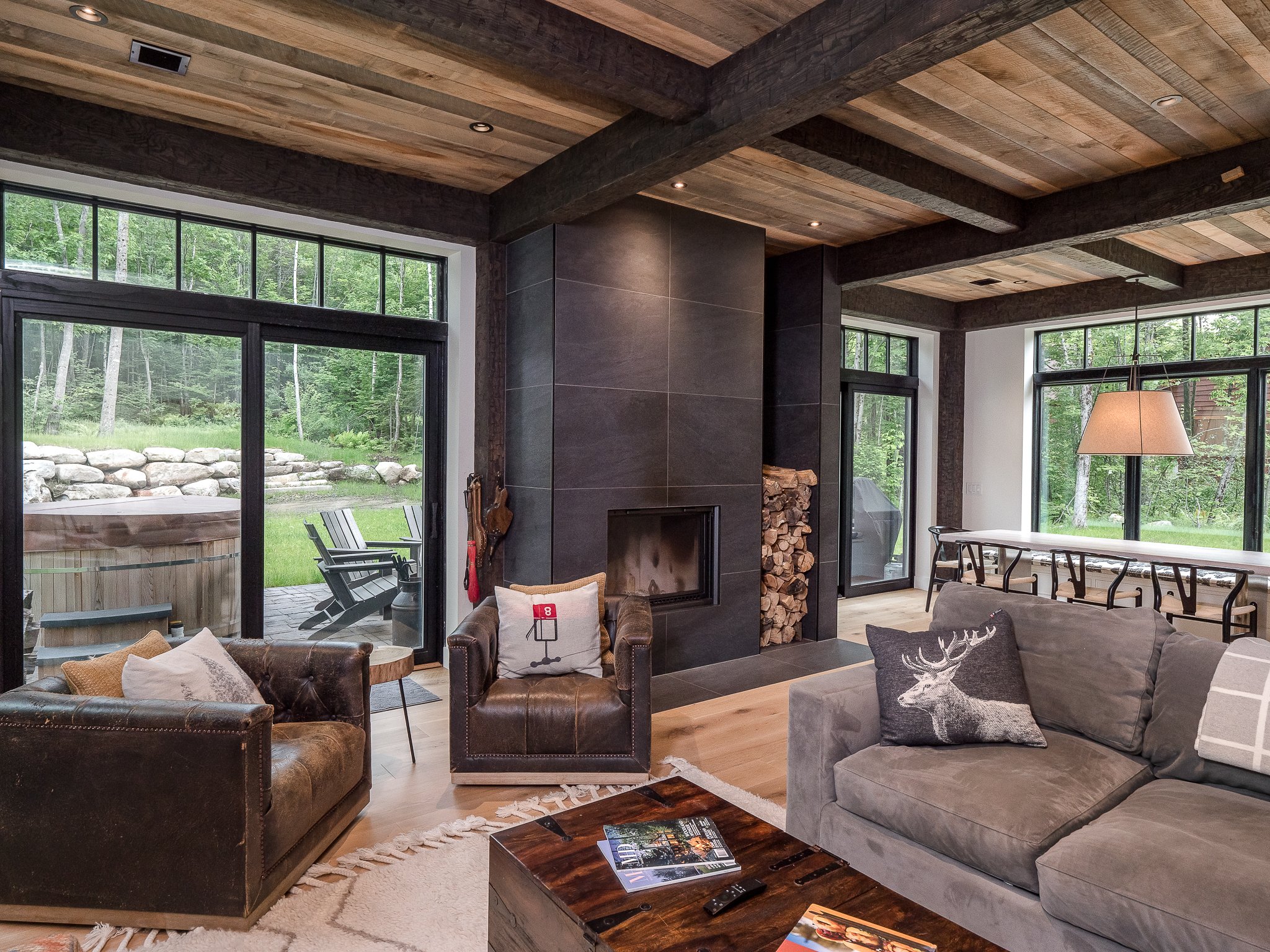 Luxury cabin interior with rustic decor, featuring a cozy seating area, large windows, and a modern fireplace. The room includes leather armchairs, a grey sofa with decorative pillows, wooden ceiling beams, and a view of the outdoor patio and forest.