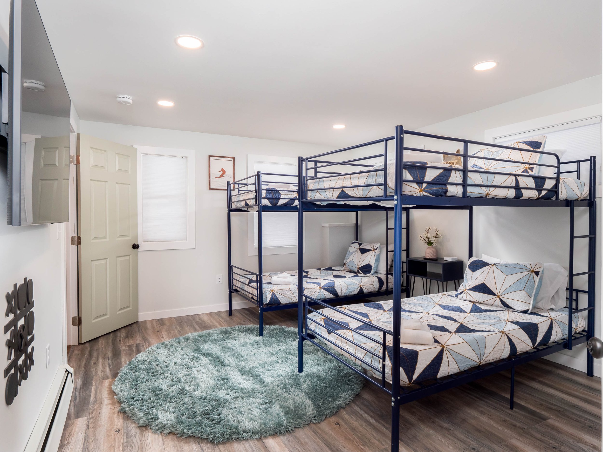 A room with three bunk beds with geometric patterned bedding, a light green circular rug, a small black side table with a flower vase, a wall-mounted TV, and framed artwork, in a well-lit space with wood flooring.