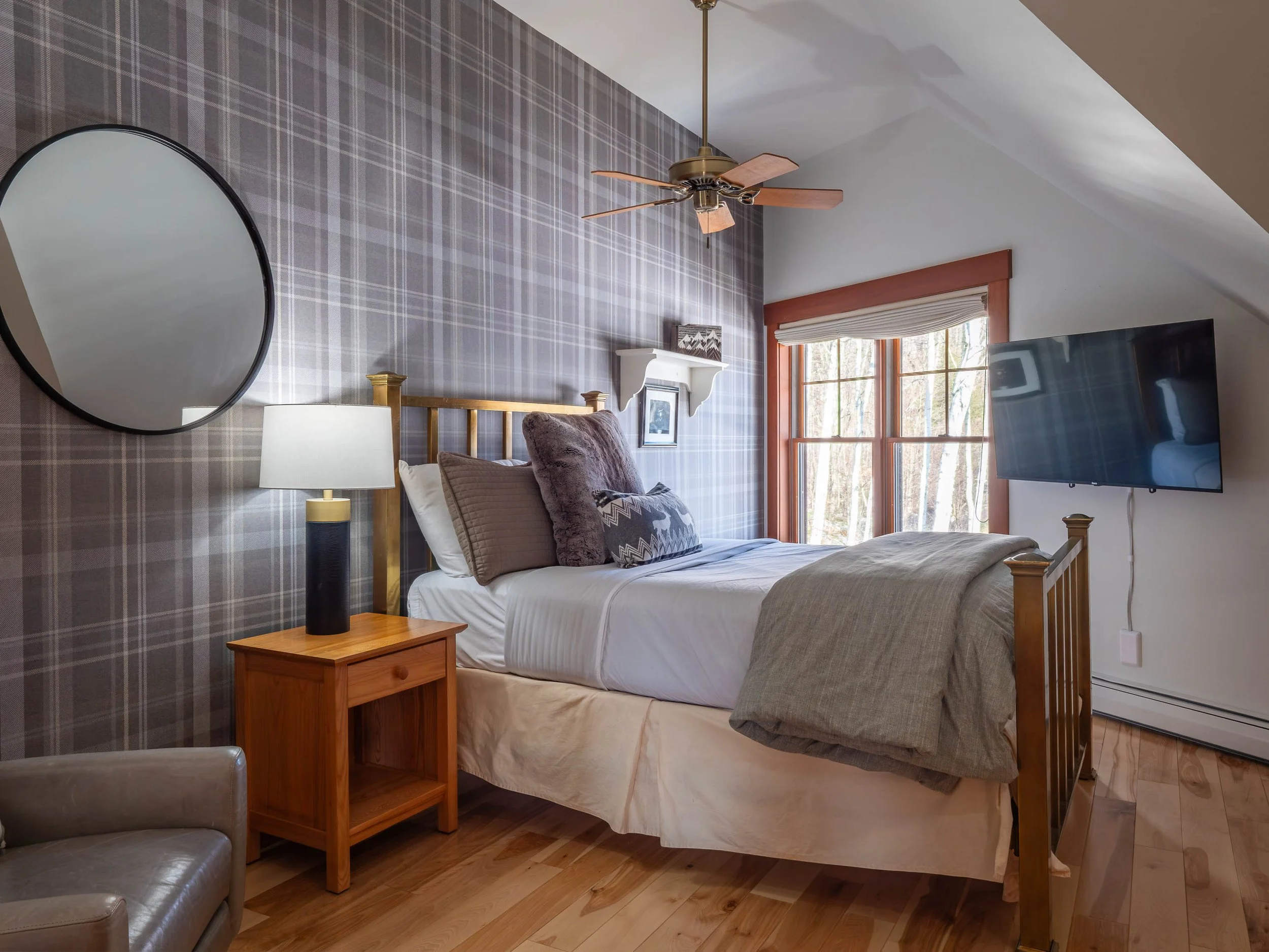 A bedroom with a double bed, gray and brown pillows, a wooden nightstand with a lamp, a wall mirror, and a window with red framing. A flat-screen TV is mounted on the wall to the right, under a ceiling fan.