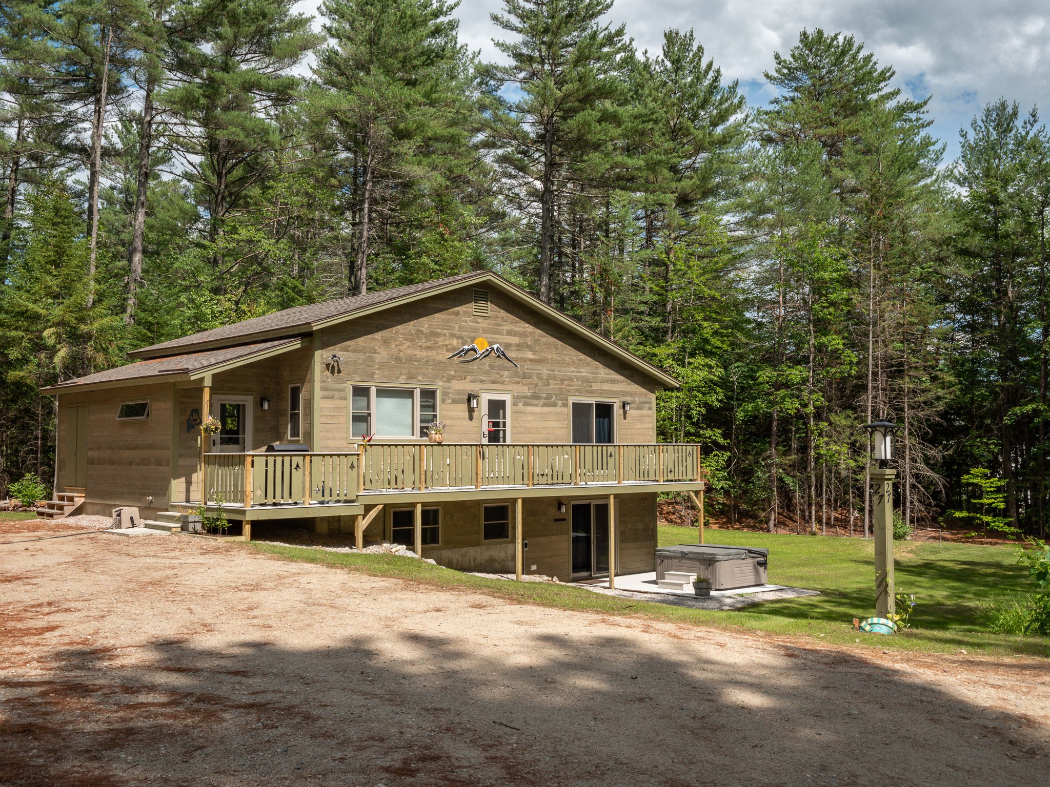 A two-story wooden cabin surrounded by tall pine trees with a gravel driveway and a deck. The house has various windows and a ground floor entrance. A hot tub is located outside on a patio area near the cabin.