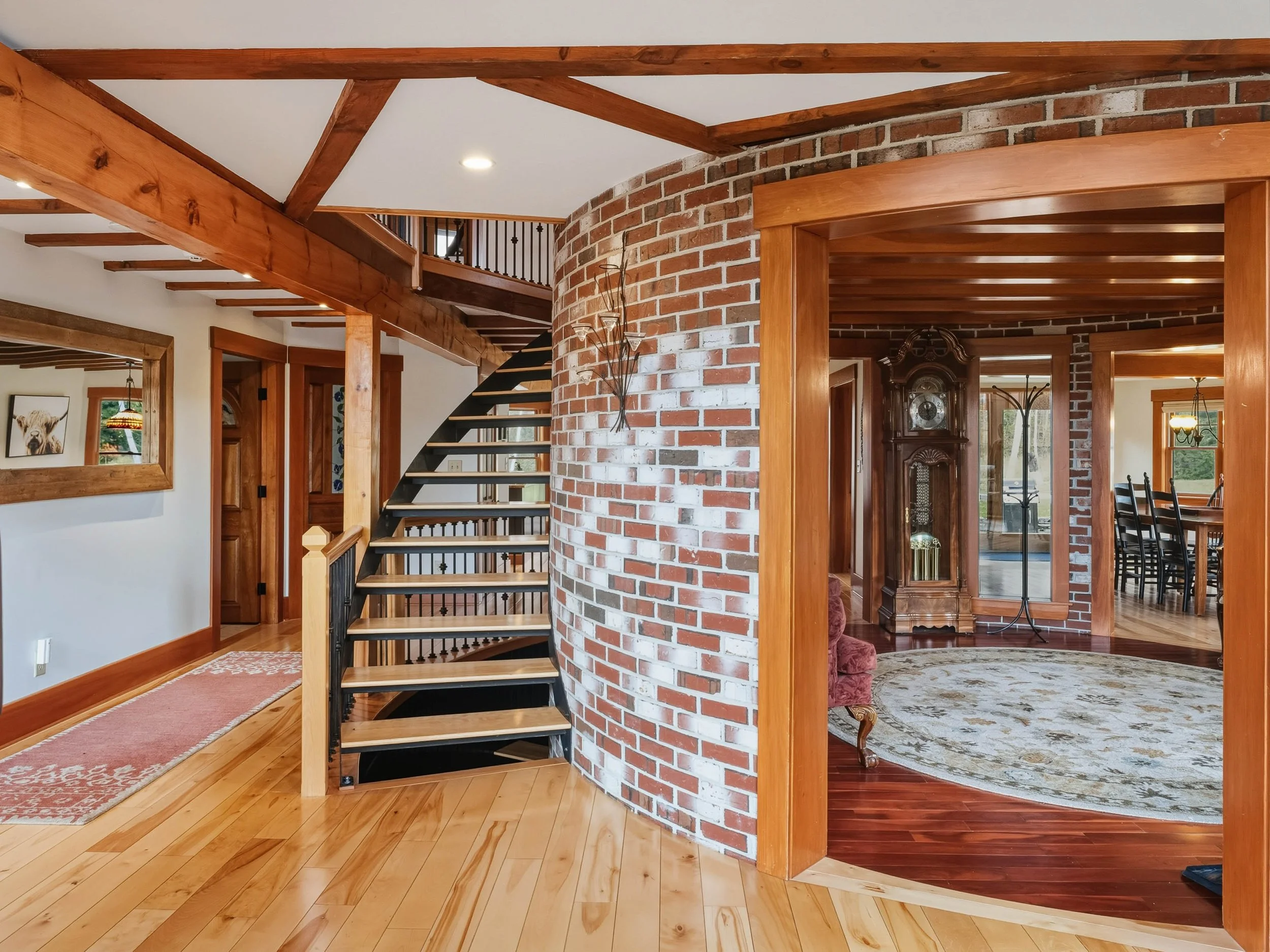 Interior view of a house with wooden floors, a curved brick wall, and a staircase with wooden treads and black risers. There is a brick wall with a metal wall sculpture, and adjacent rooms with wooden trim, furniture, and decor.