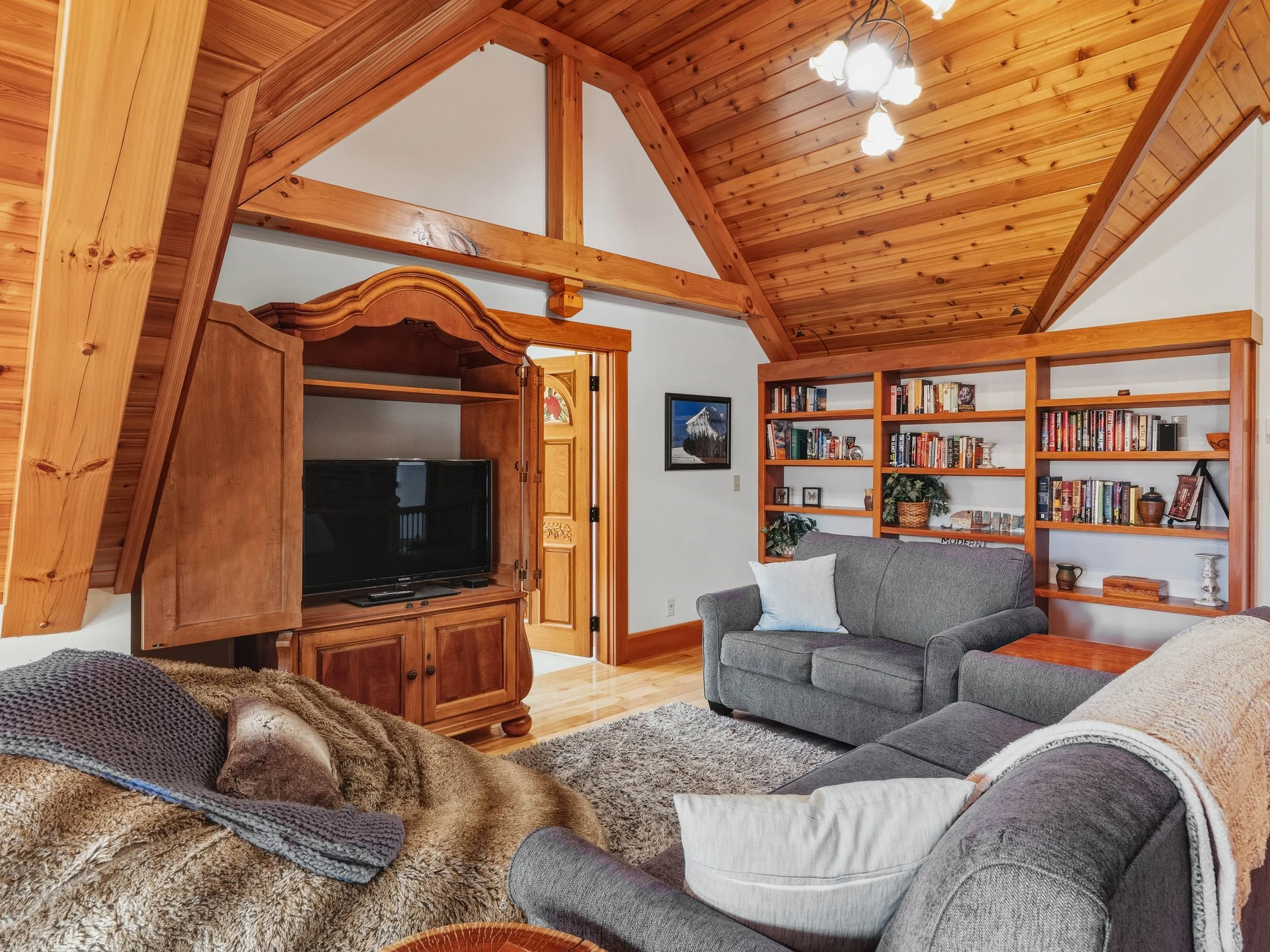Cozy living room with wood paneling on the ceiling, a TV in a wooden cabinet, gray couches, and a wooden bookshelf filled with books and decorative items.