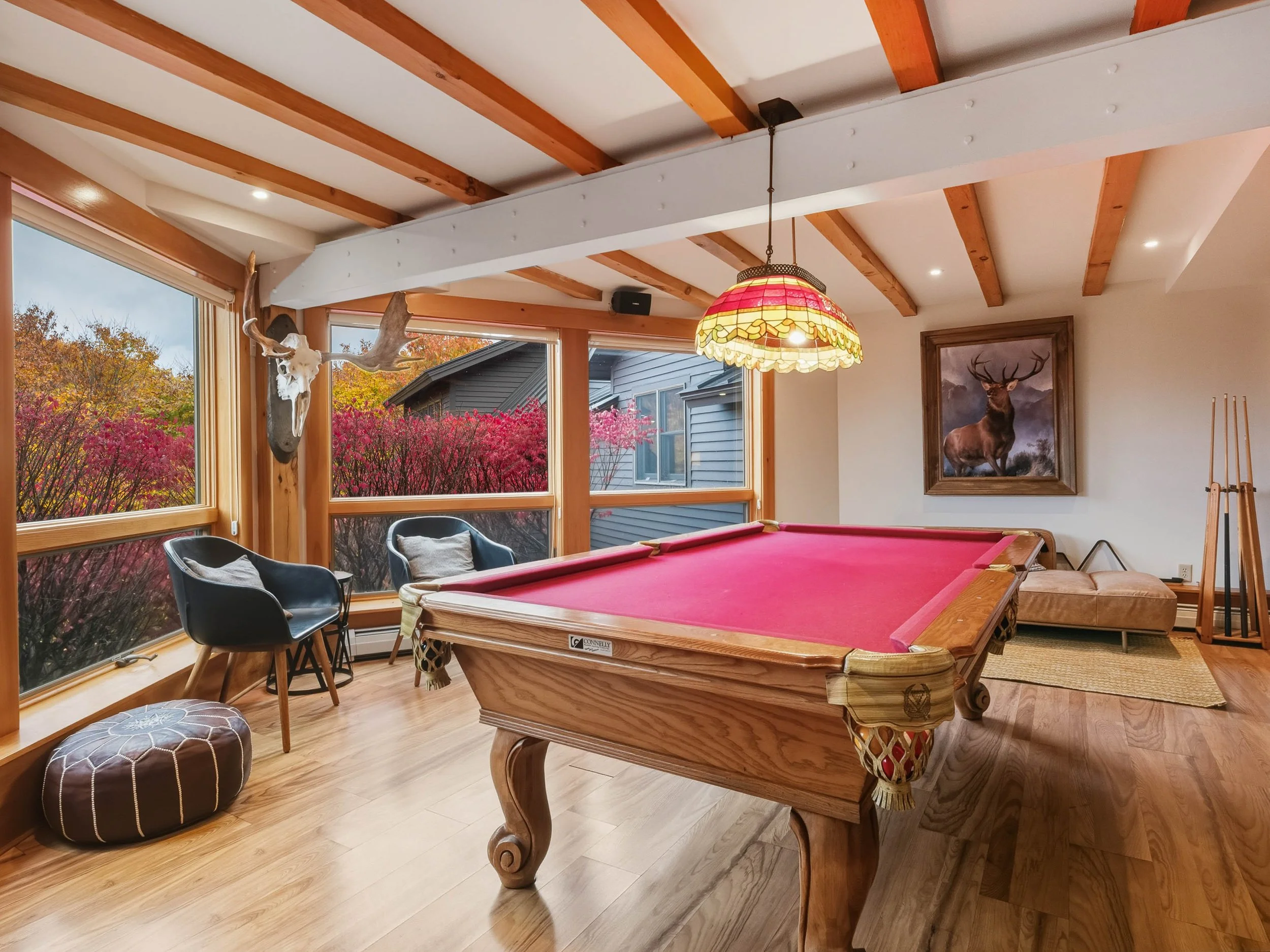 A cozy game room with a pink pool table, a stained glass hanging lamp, two black chairs, a pouf, large windows showing autumn foliage, a deer skull with antlers on the wall, and a framed painting of a buck hanging above a beige bench.