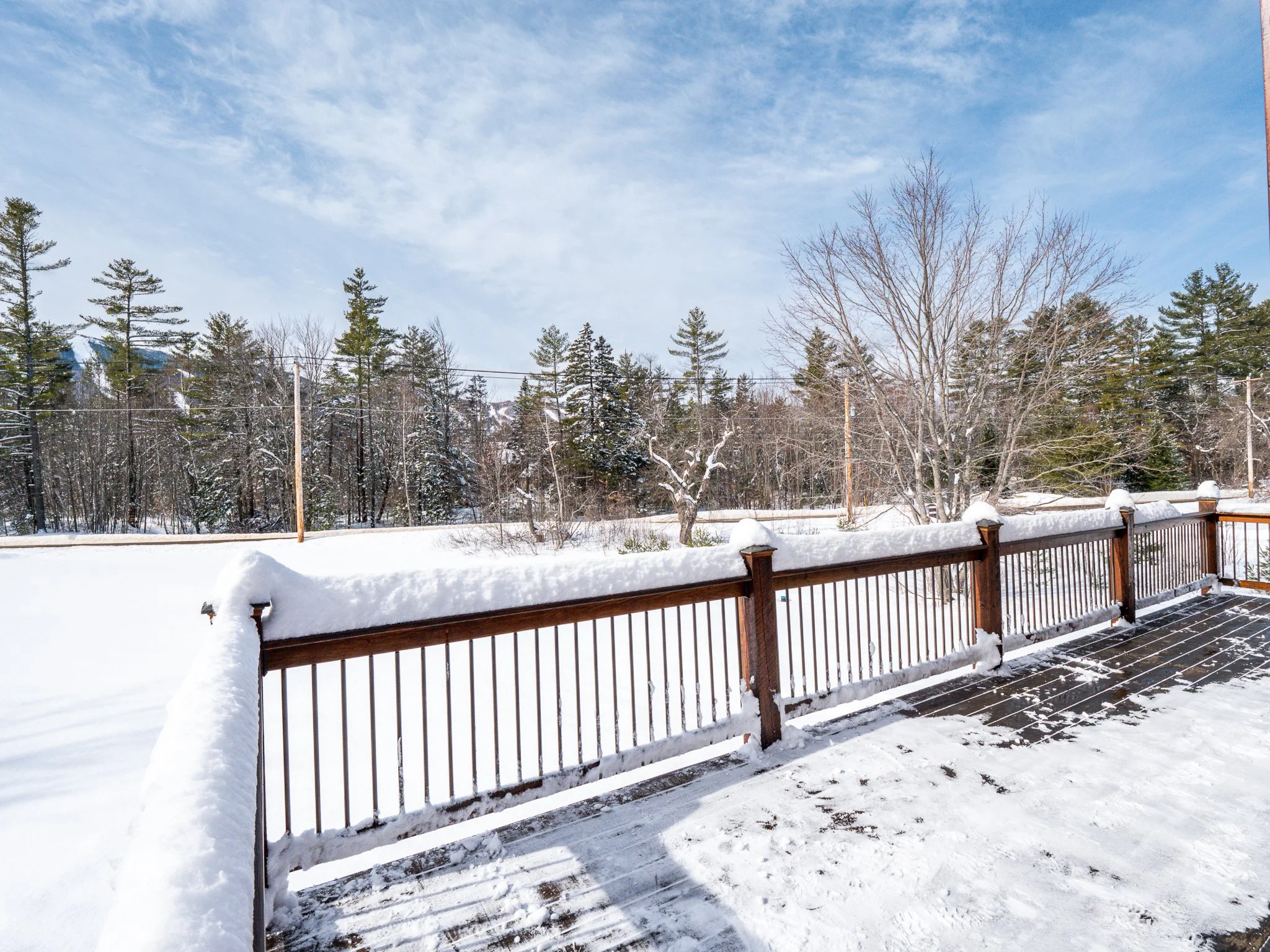 Snow-covered wooden deck with railing overlooking snowy landscape with trees and clear sky