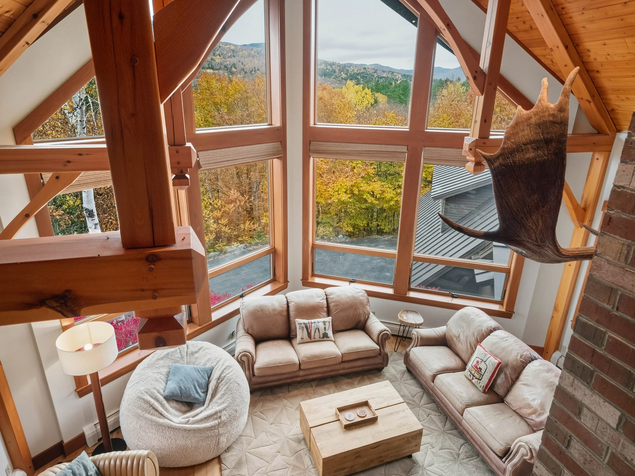 Living room with large windows showcasing an autumn landscape, wooden beams, a beige couch, a pink leather sofa with decorative pillows, a bean bag chair, a wooden coffee table, a side table, and a mounted animal hide on the wall.