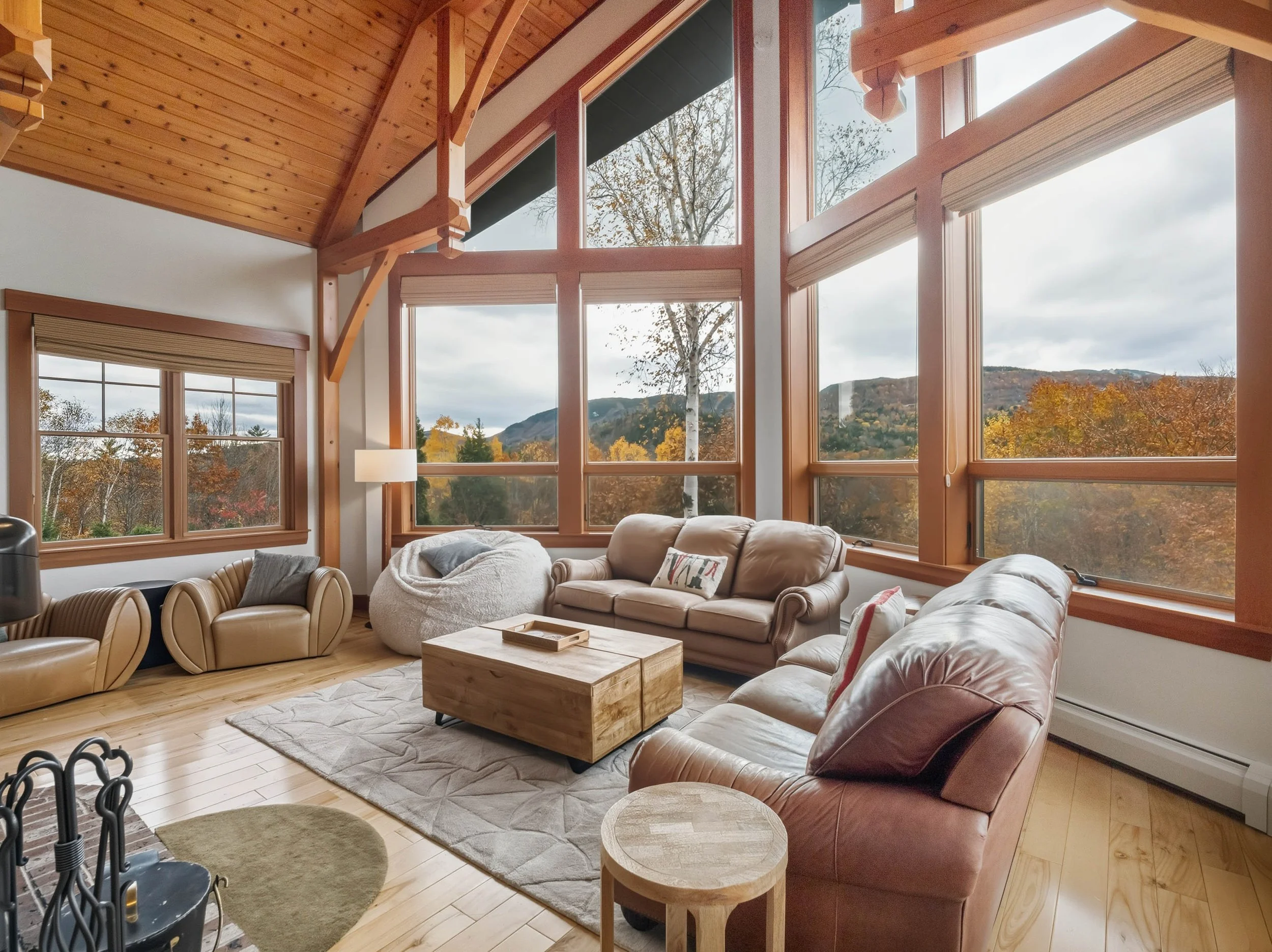 Sunlit living room with large wood-framed windows viewing fall trees and mountains, decorated with leather sofas, armchair, big bean bag, wooden coffee table, area rug, and nested pillows.