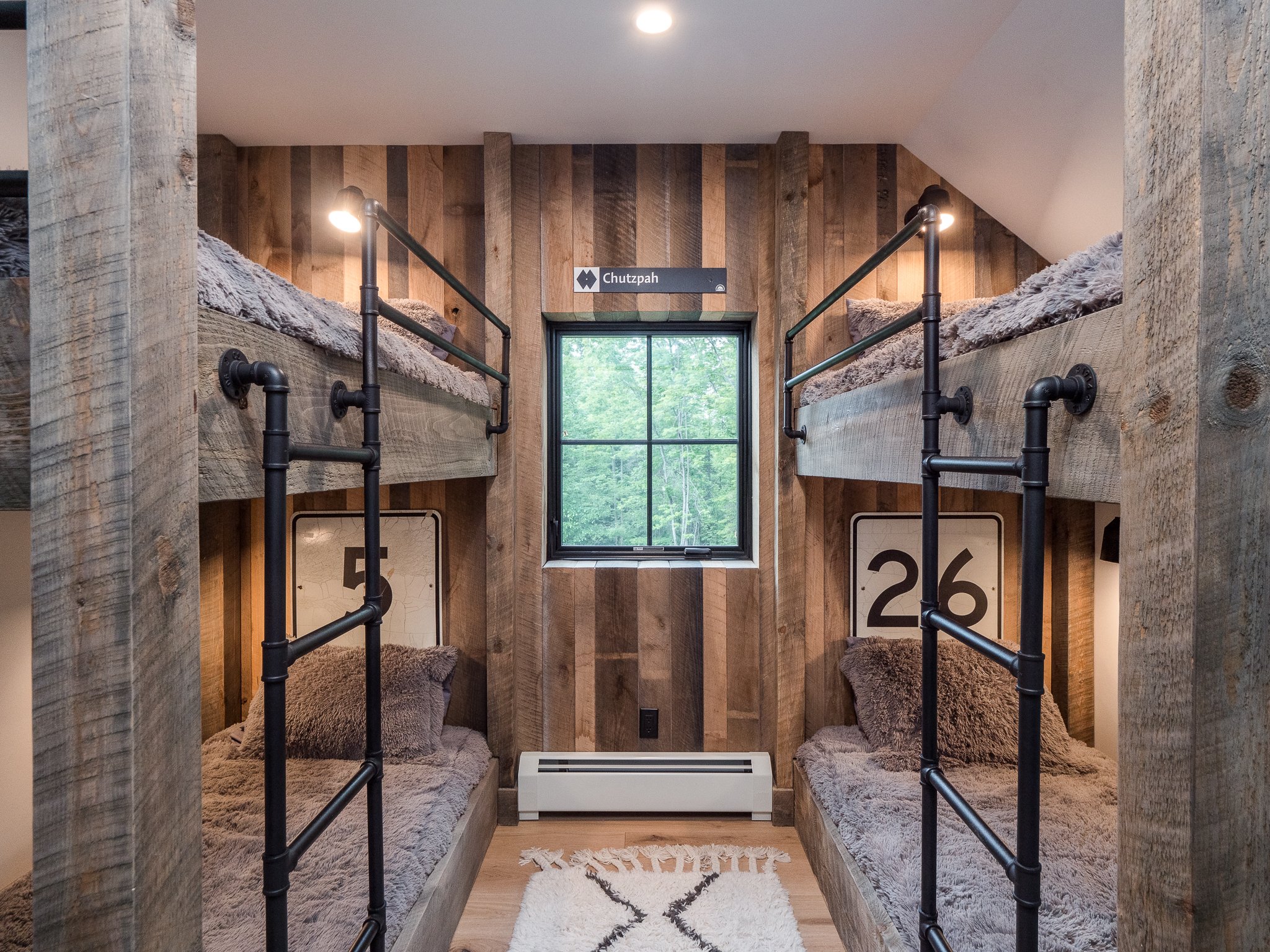 Rustic bunk room with wooden walls, two sets of bunk beds, and metal ladders, featuring cozy bedding and a central window.