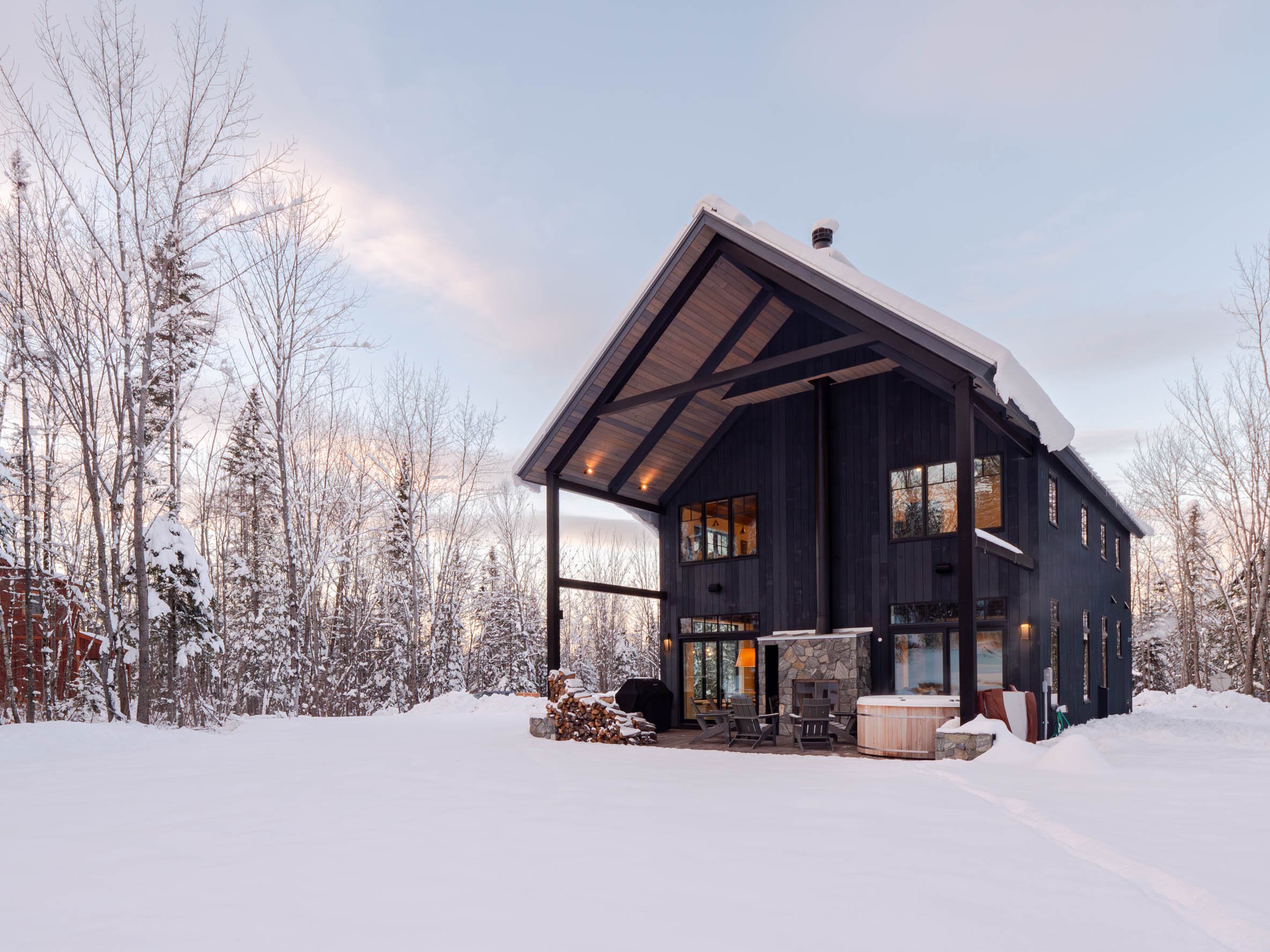 Modern two-story black house with large windows, surrounded by snow-covered trees and landscape, featuring an outdoor seating area and hot tub.