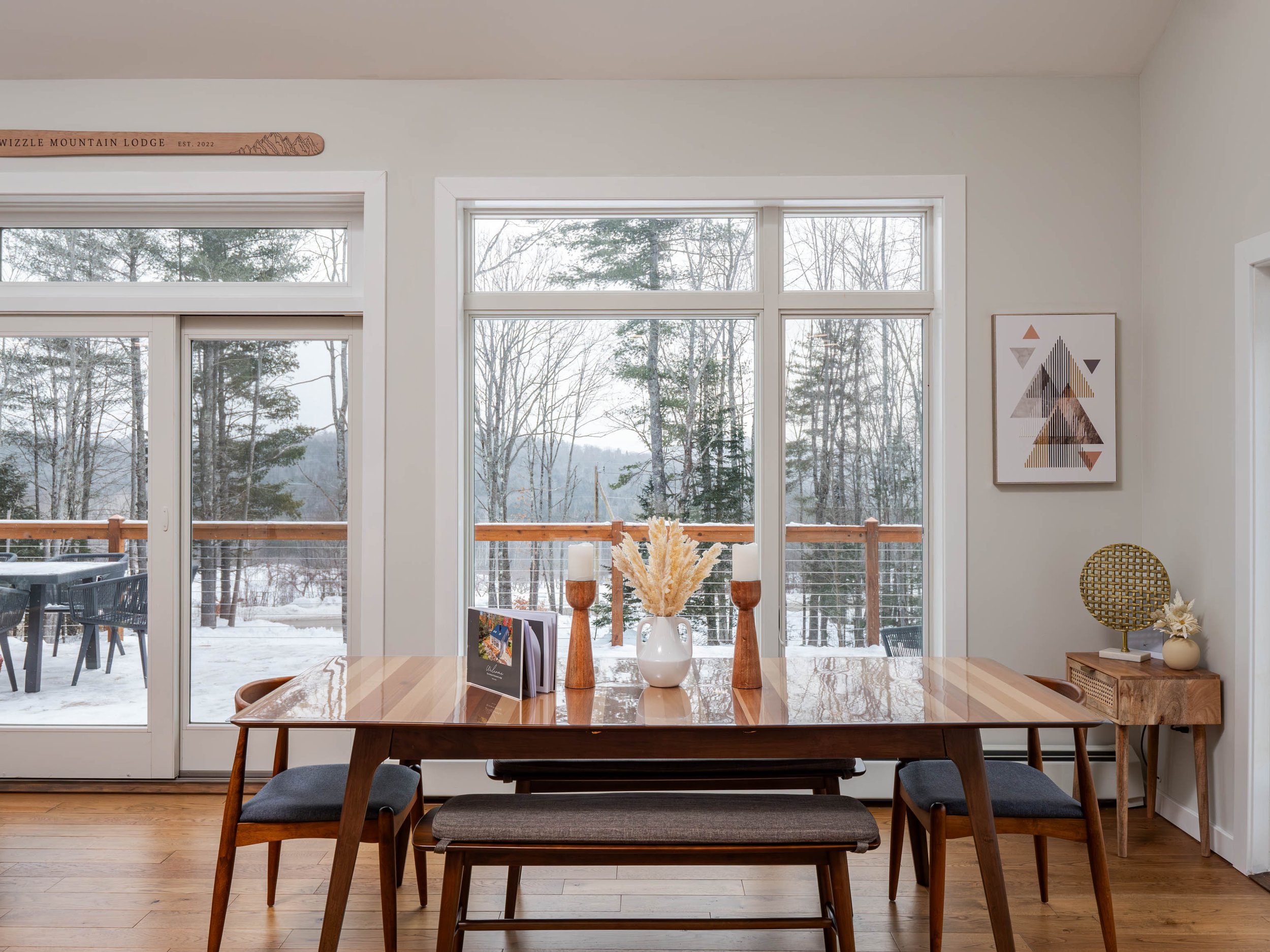 Dining room with wooden table, benches, and chairs, large windows overlooking a snowy outdoor patio and woods, decor including vases and artwork.