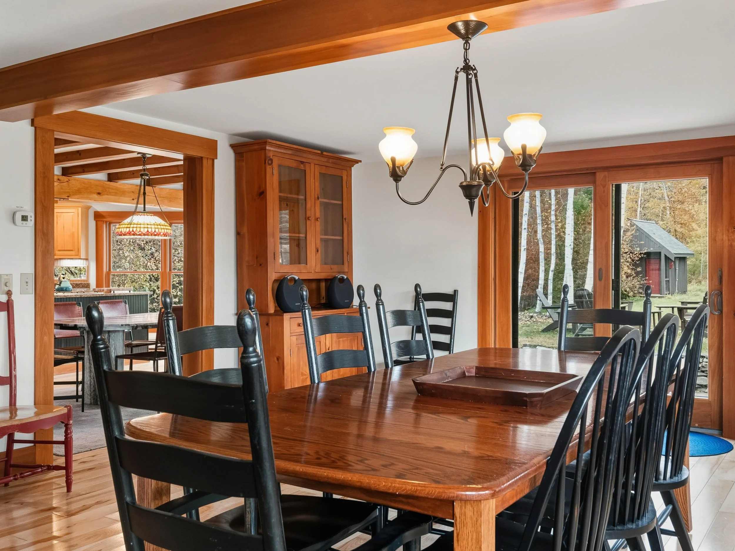A dining room with a large wooden table and black chairs. A chandelier hangs above the table, and large sliding glass doors lead to an outdoor area with a shed and trees.