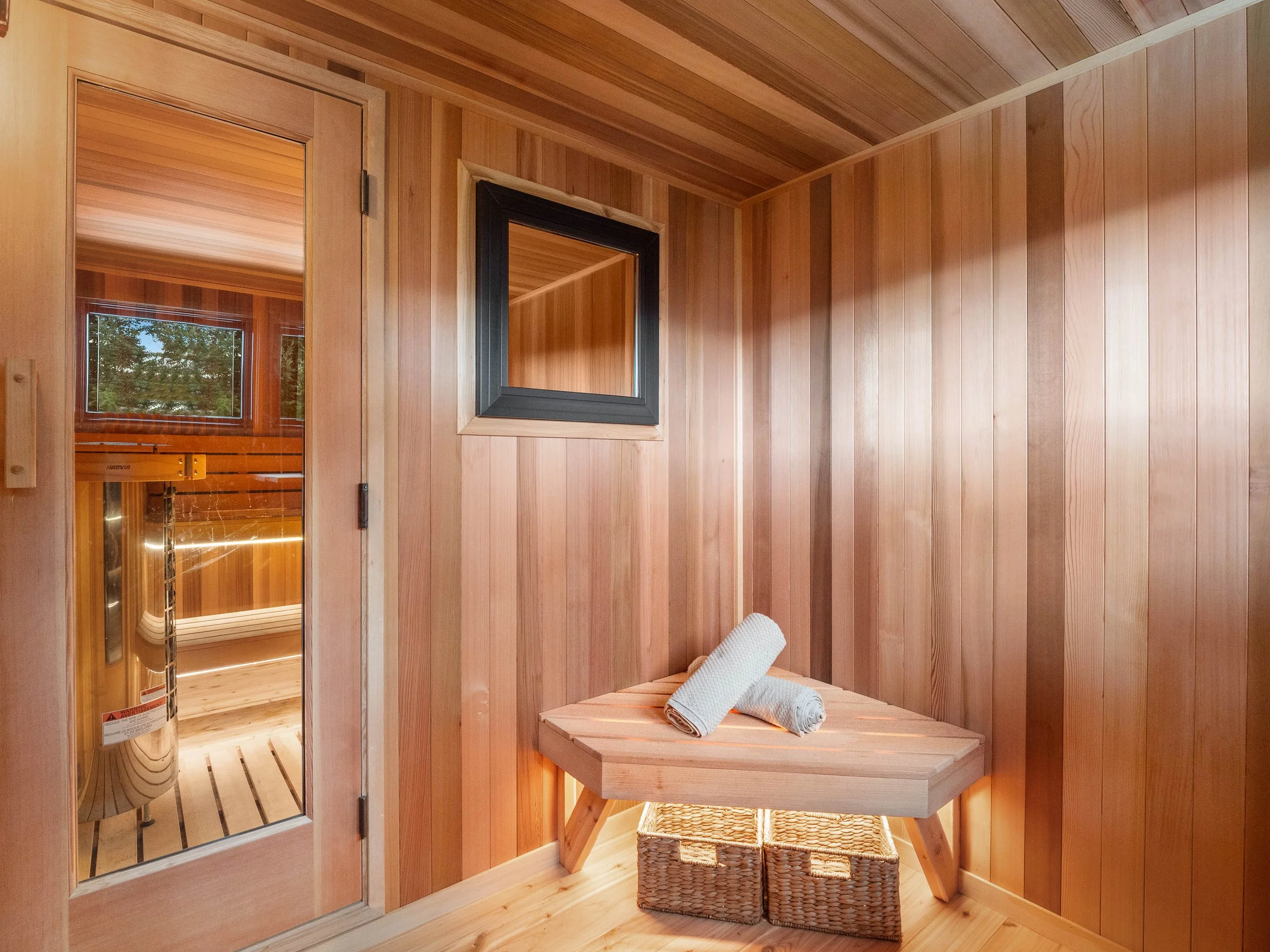 Wood-paneled sauna room with a bench, two rolled white towels, a small window, and a glass door leading to the sauna interior.