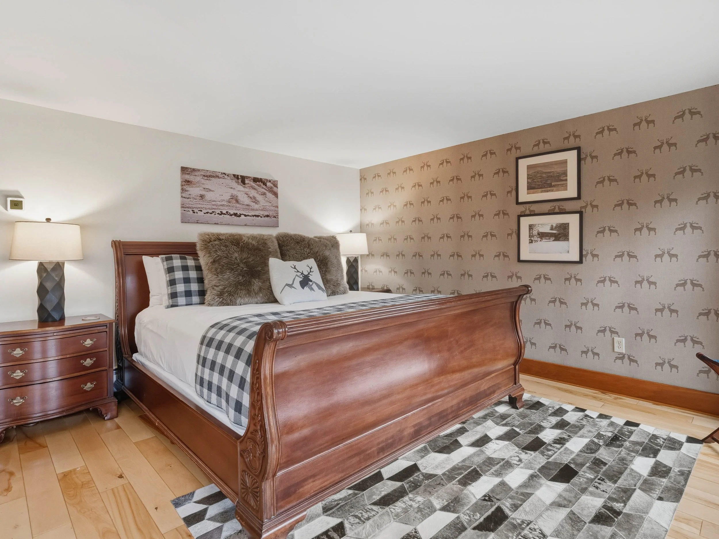 A bedroom with a wooden bed frame, plaid and furry pillows, a patterned wallpaper with reindeer, and black and white framed pictures on the wall.