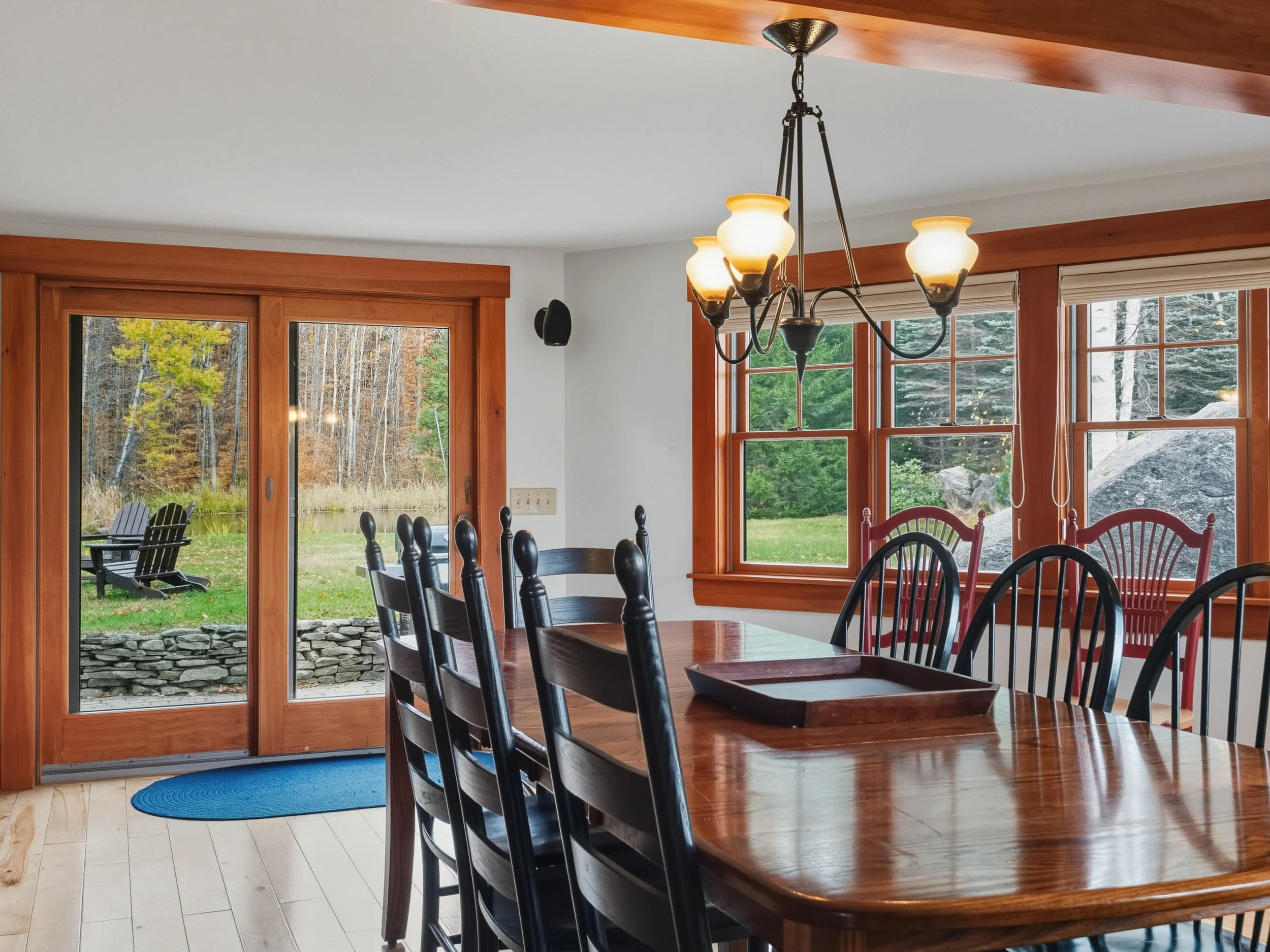 Dining room with wooden table and matching chairs, large windows showing outdoor greenery, sliding glass door leading to a patio with Adirondack chairs, chandelier hanging from ceiling, wood trim accents, hardwood floor, and natural light.