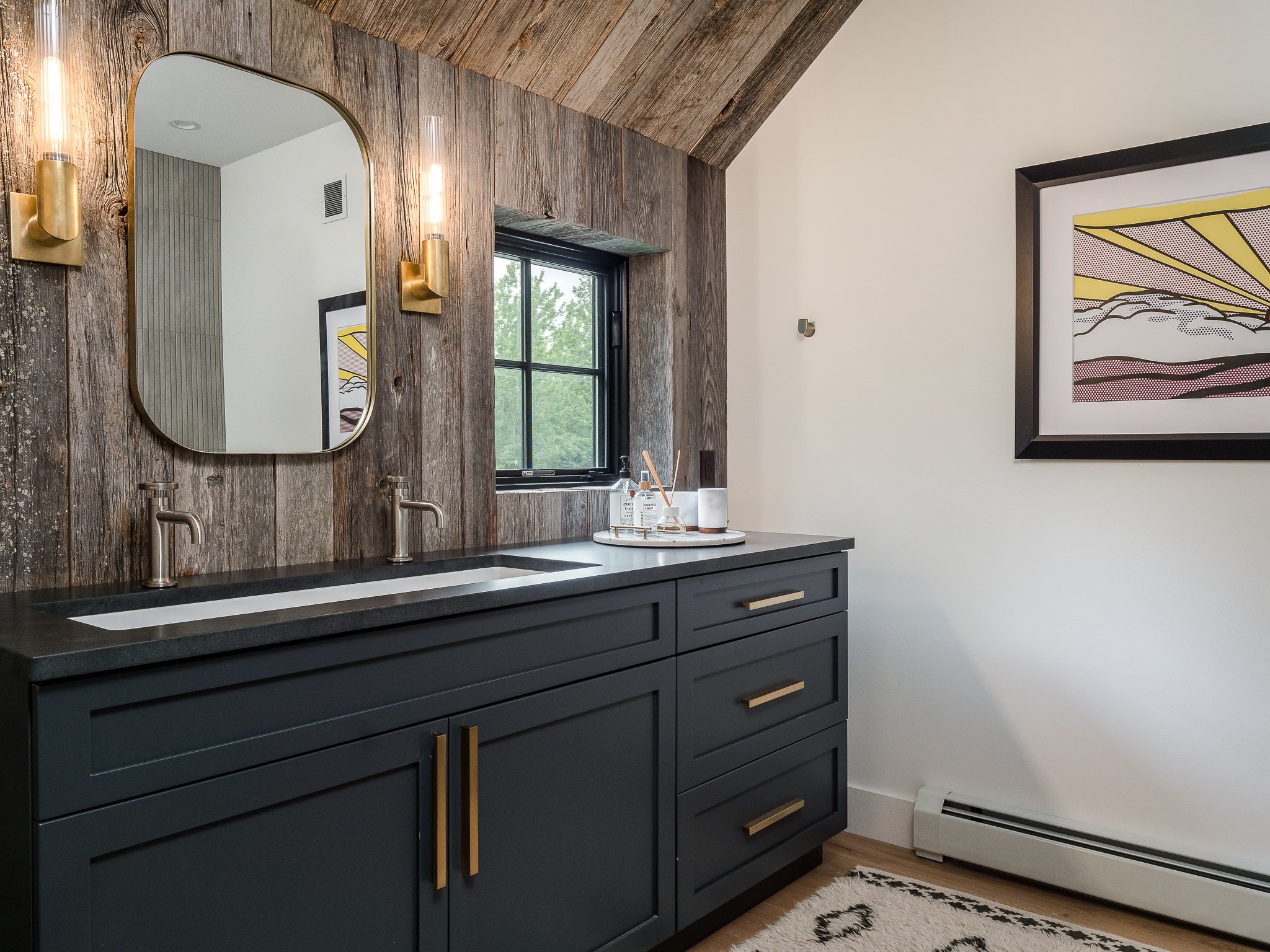 Modern bathroom with dark vanity and double sinks, rustic wood wall, oval mirror, wall sconces, framed artwork, and window.