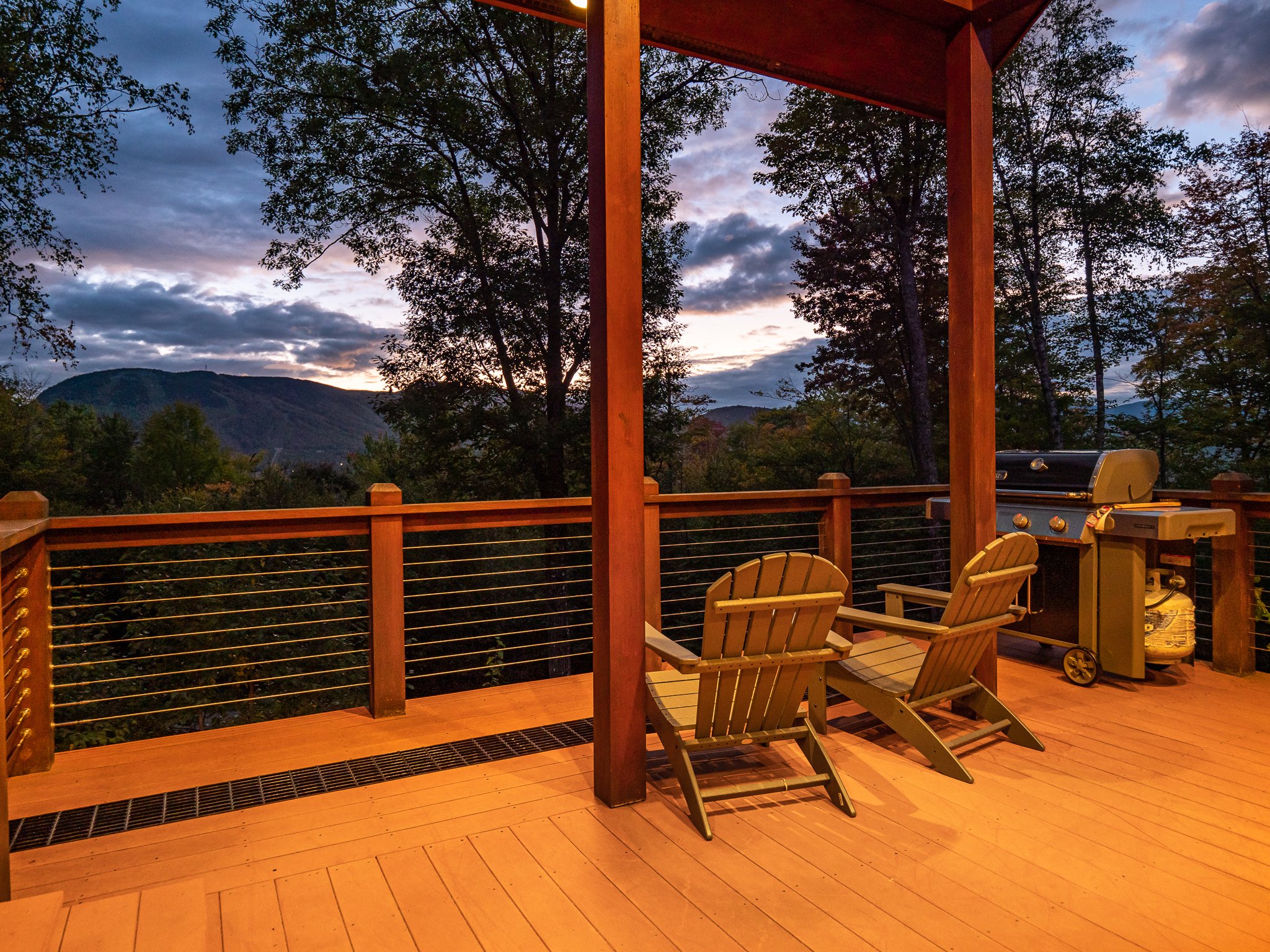 Wooden deck with Adirondack chairs, a grill, and a view of mountains at sunset.