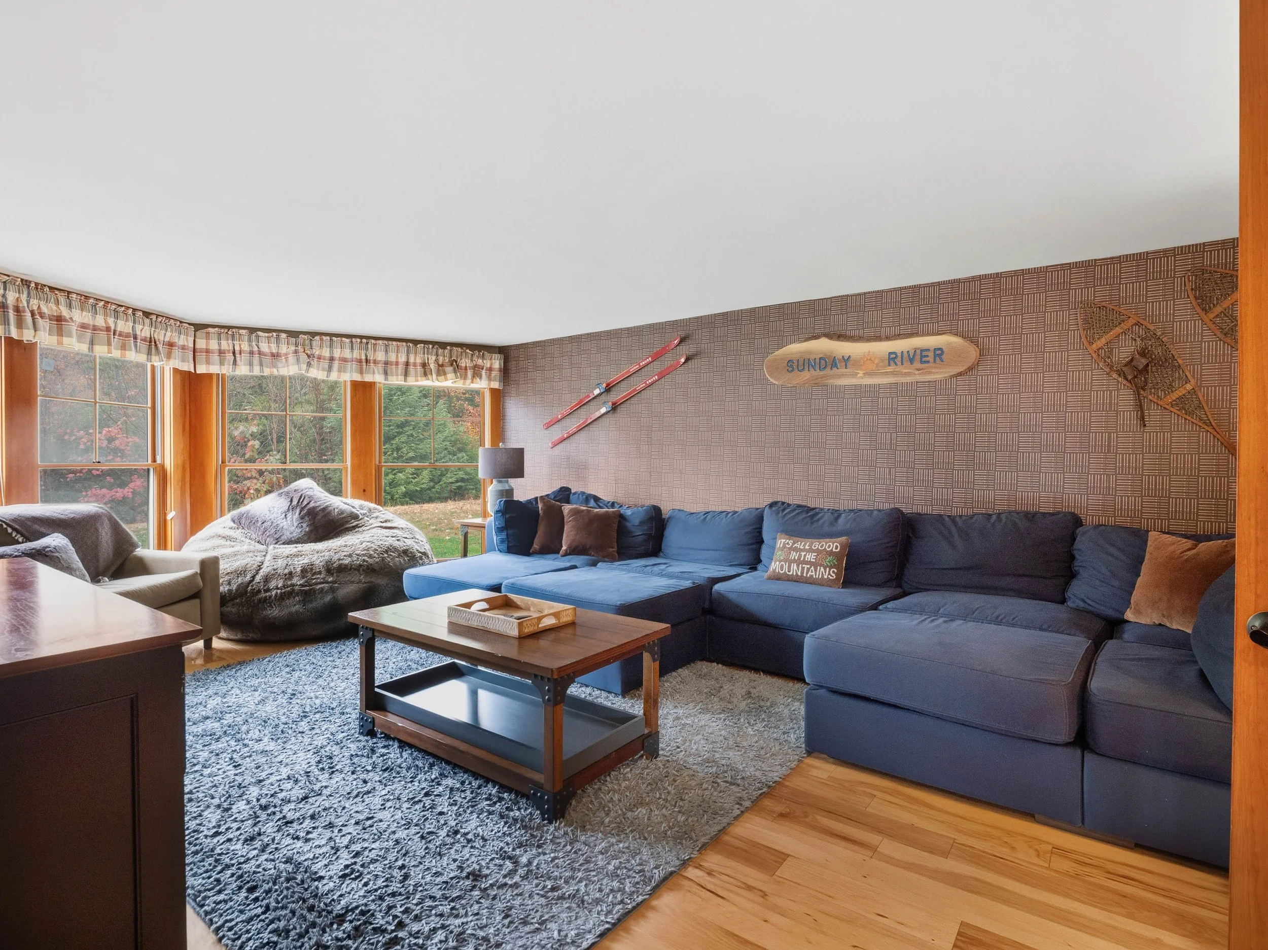 Living room with large windows, blue sectional sofa, gray armchair, mountain-themed decor, skis, and a rug.