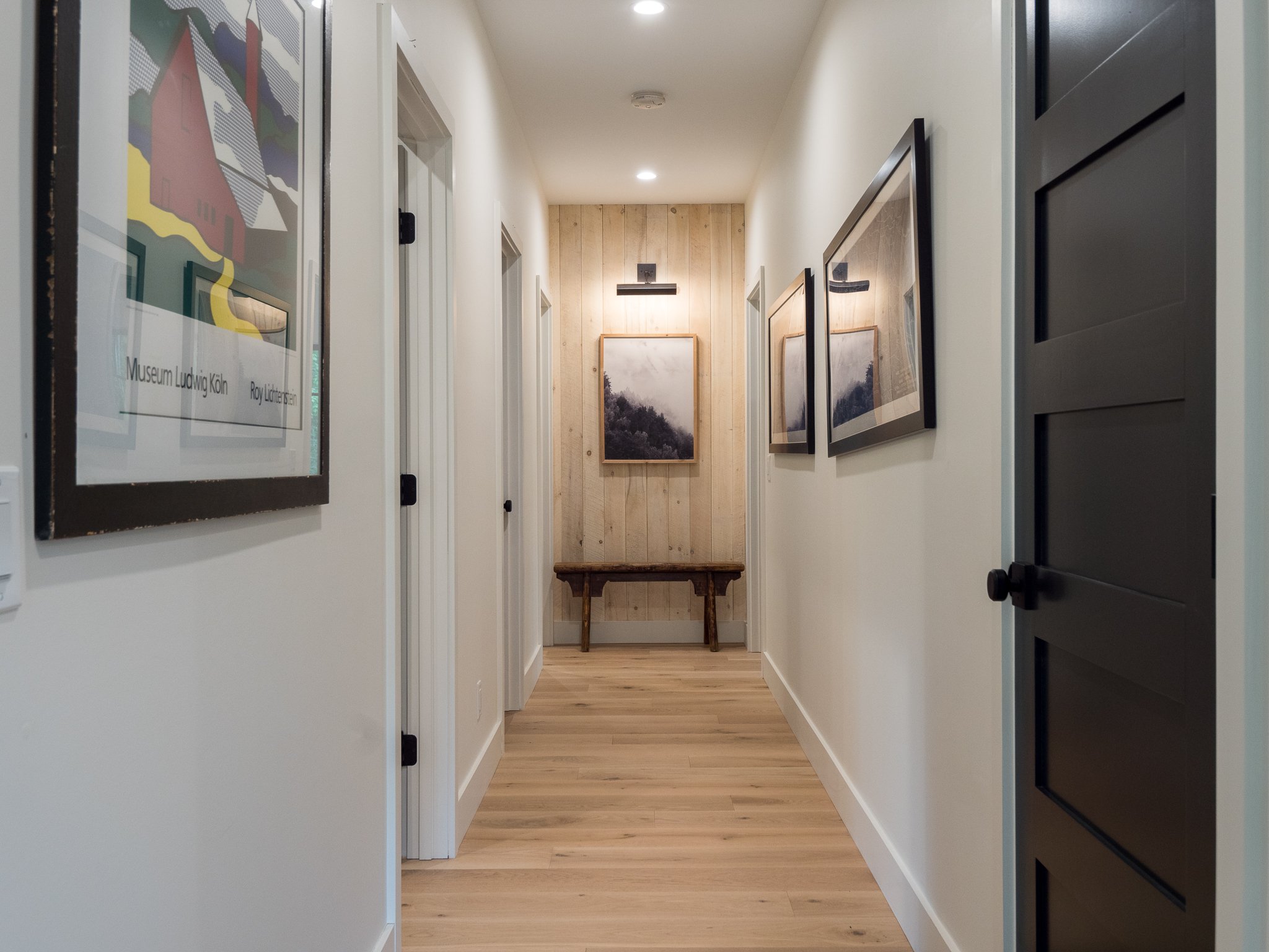 Hallway with light wood flooring, white walls, black framed artworks on walls, black doors, and a small wooden bench at the end.