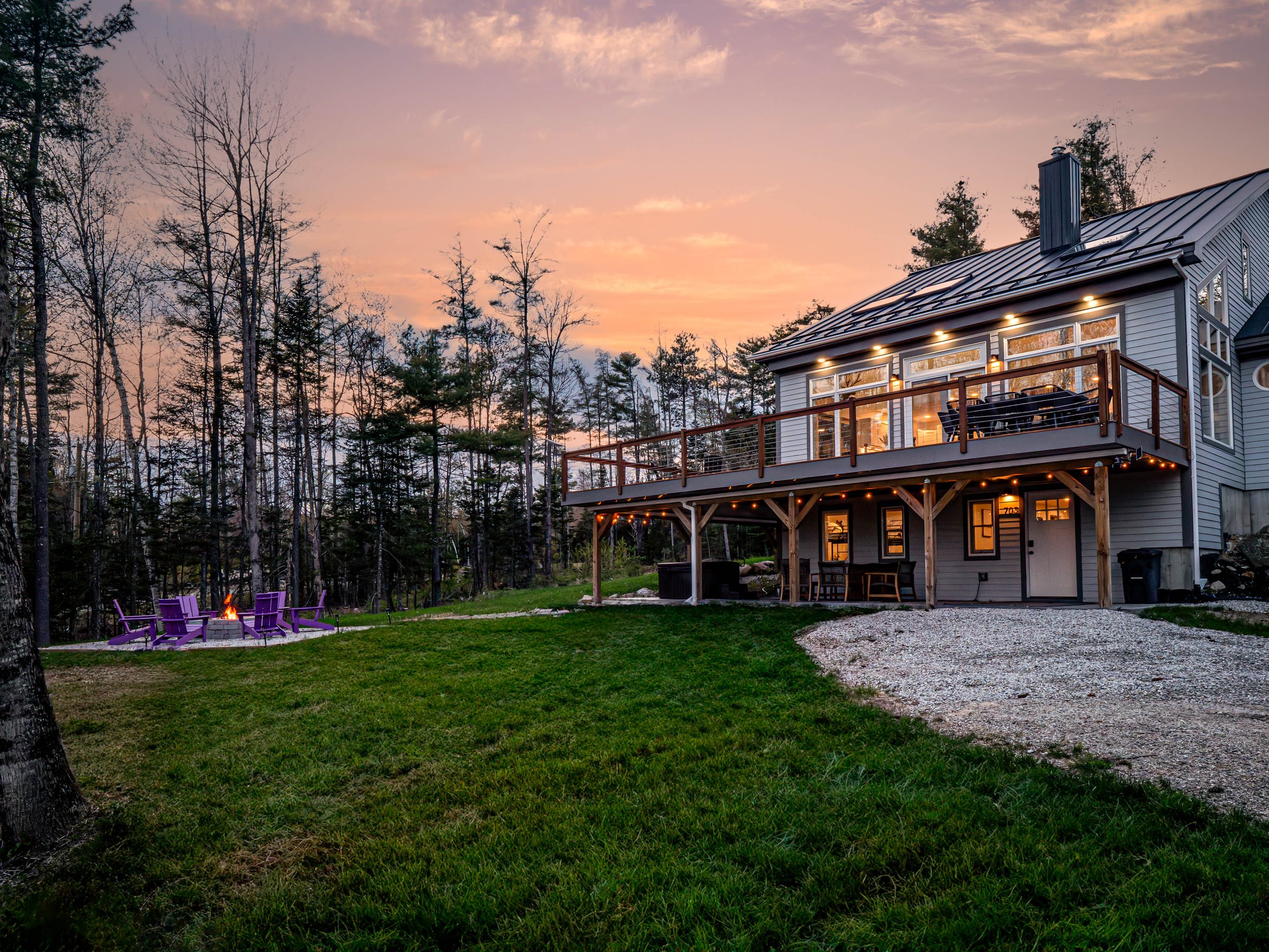 A two-story house with a large balcony illuminated at sunset, surrounded by a grassy yard with a fire pit and purple chairs, with a backdrop of trees and a colorful sky.
