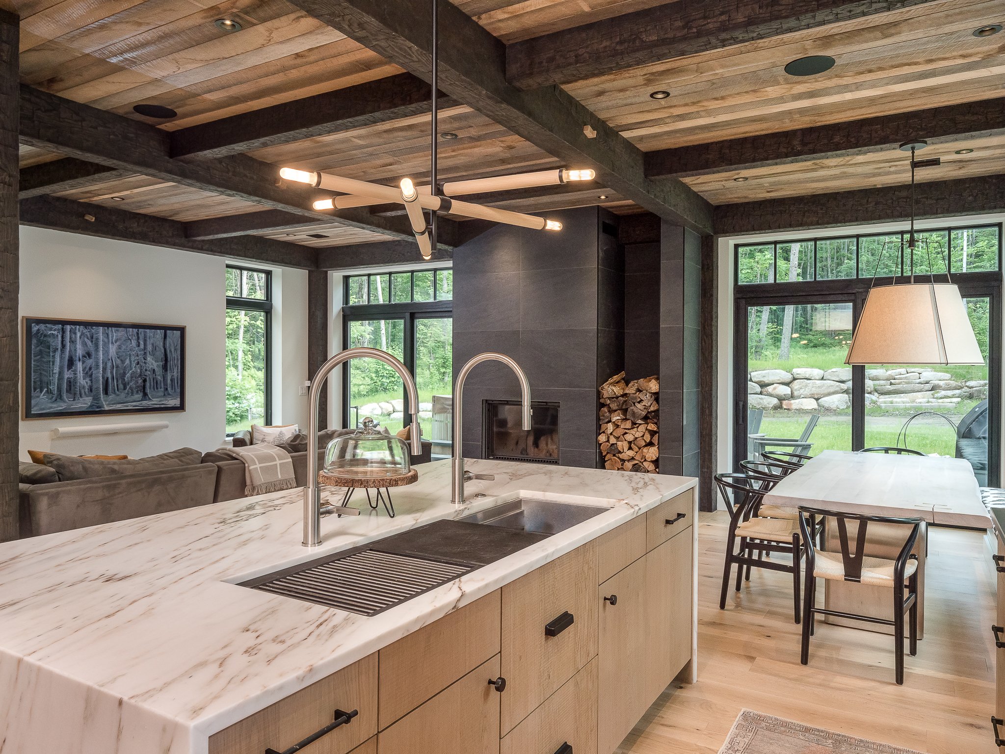 Modern interior of a living space featuring a kitchen island, wooden ceiling beams, large windows, a dining table with chairs, and a fireplace with stacked firewood.
