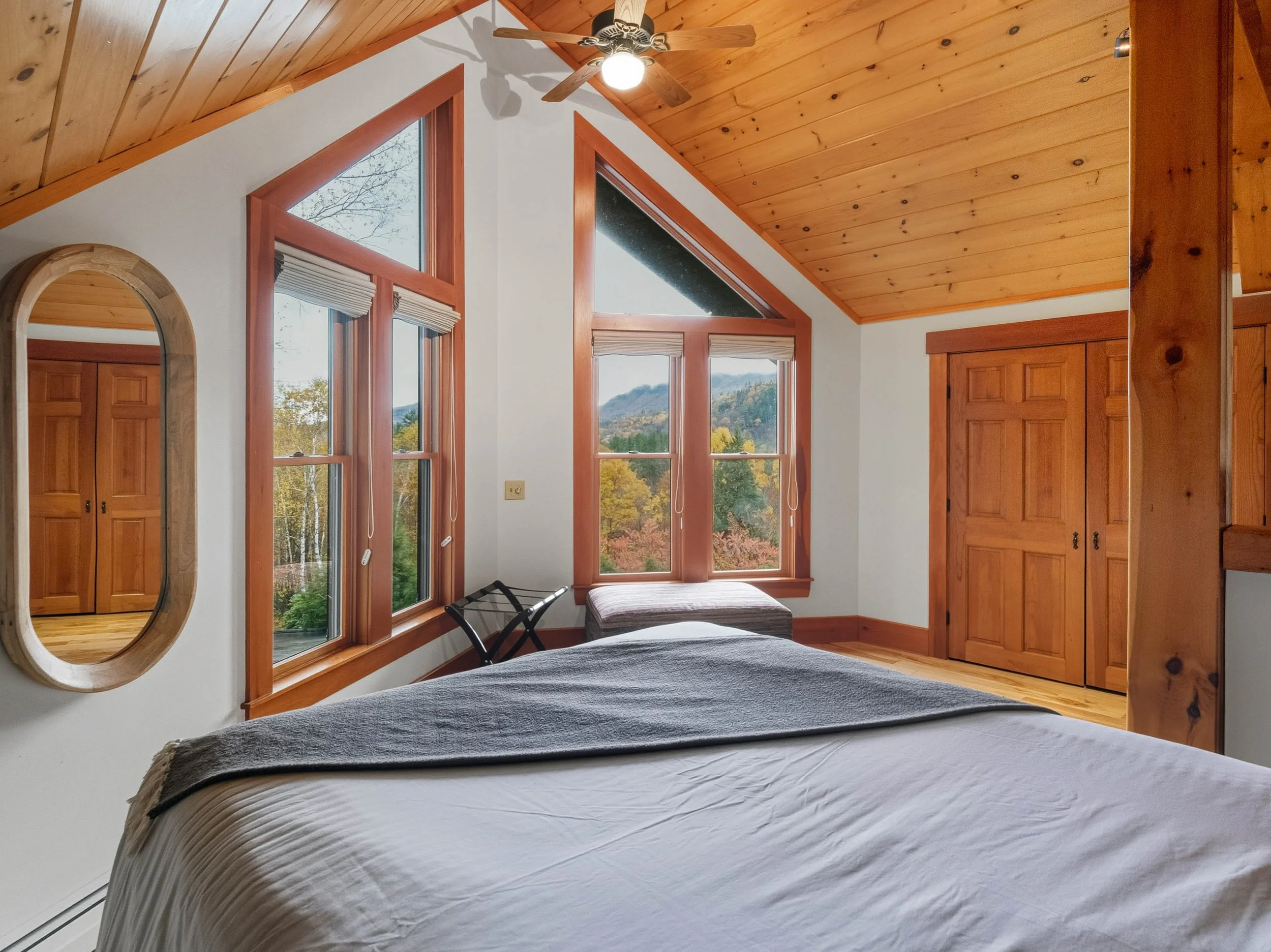 A cozy bedroom with wooden-paneled sloped ceiling, large windows showing trees and mountains, a bed with white sheets and a gray blanket, a wooden wardrobe, an oval wall mirror, a ceiling fan, and a window seat