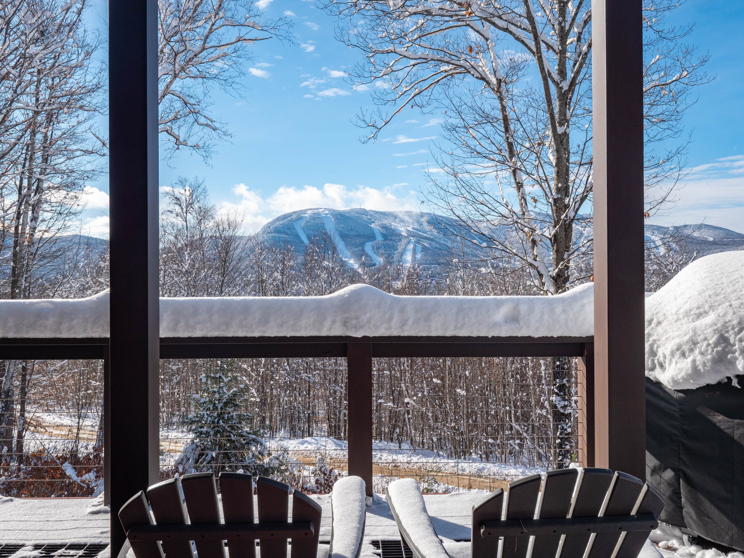 Snow-covered mountain view from a deck with Adirondack chairs and bare trees on a sunny day.