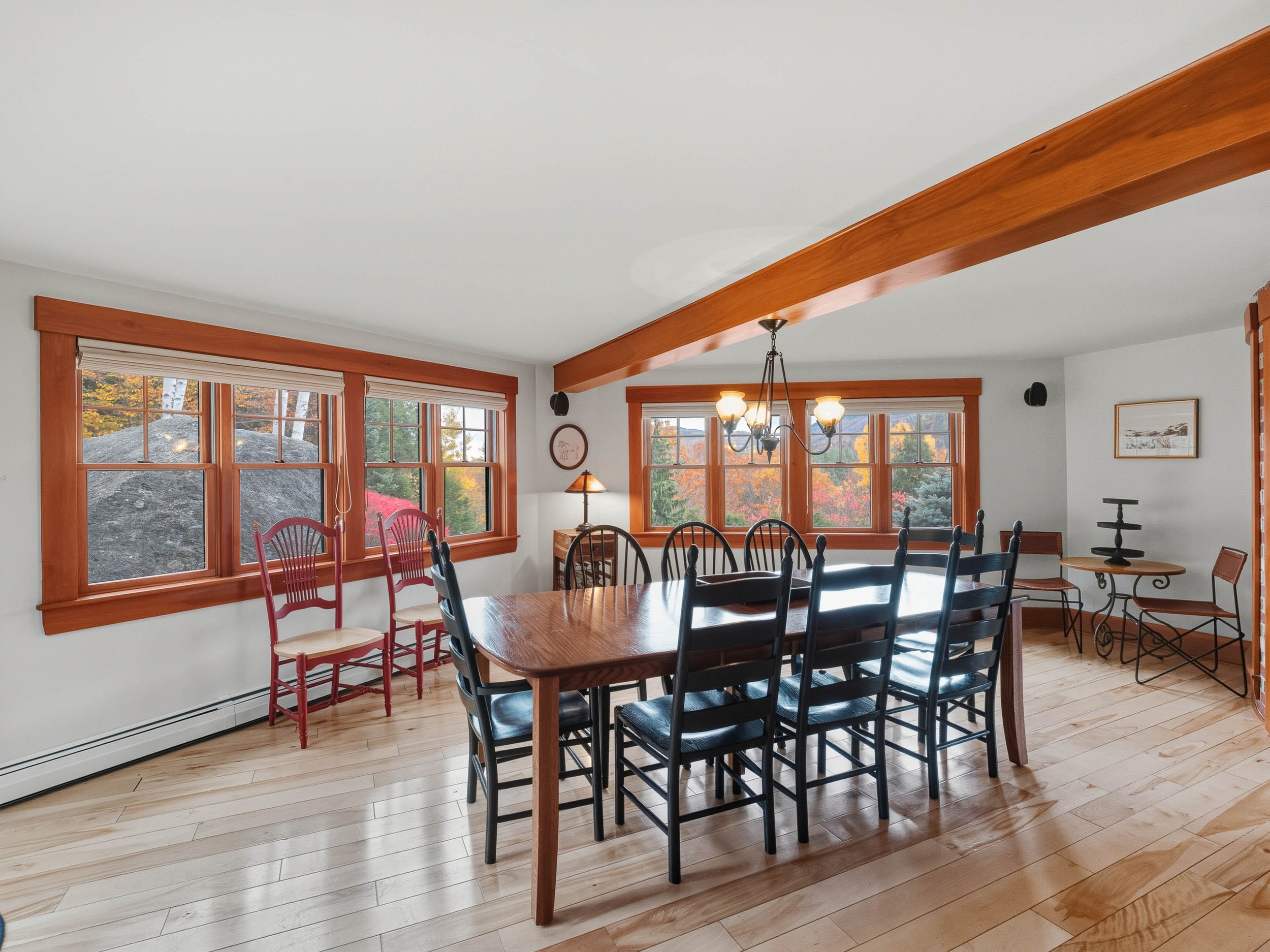 A dining room with a wooden table, black chairs, and windows showing autumn trees outside.