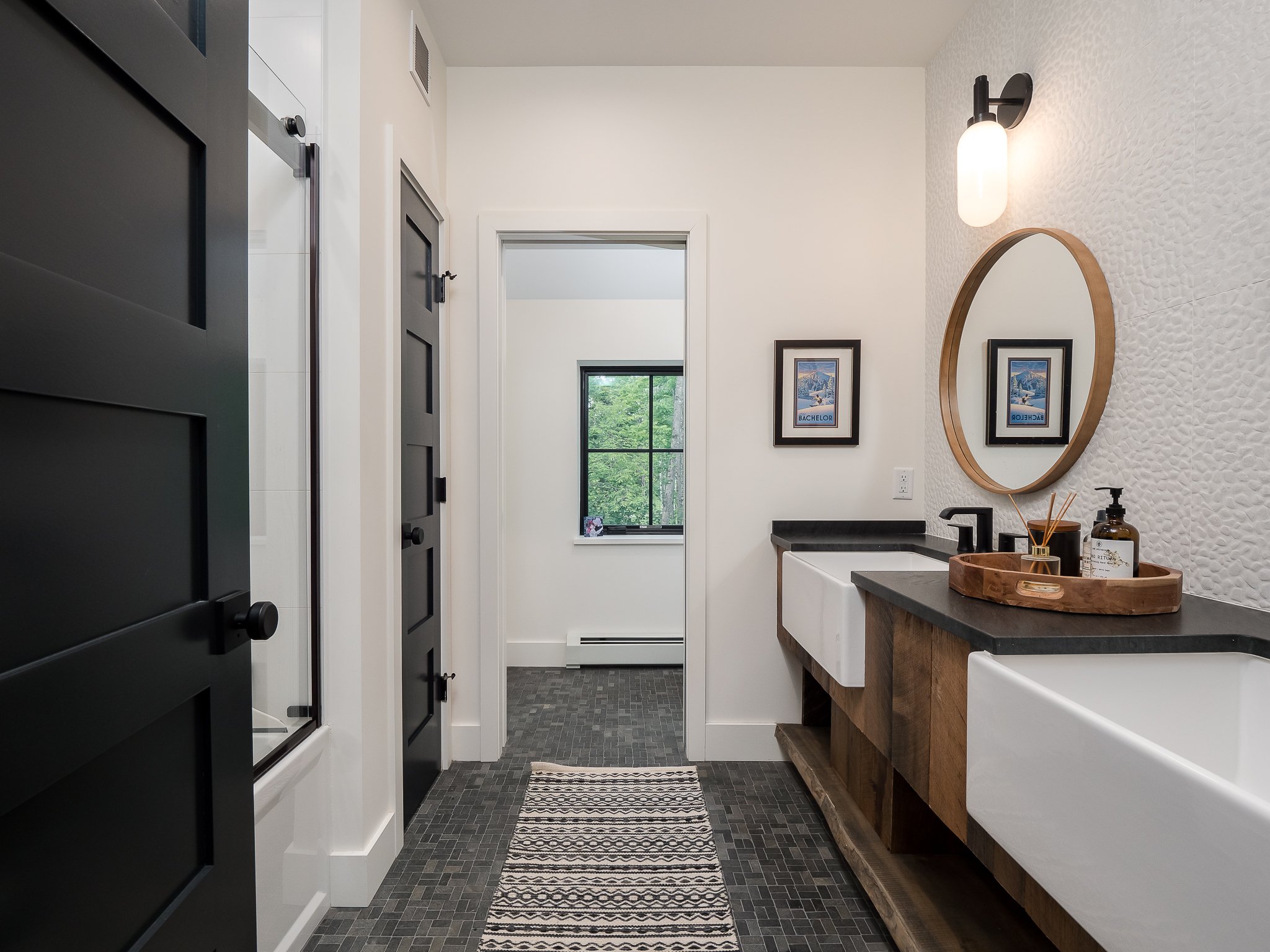 Modern bathroom with a double sink vanity, circular mirror, and dark gray tile flooring. Features two black doors, a decorative wall with textured tiles, a wall-mounted light fixture, and framed artwork. A tray with assorted toiletries sits on the co