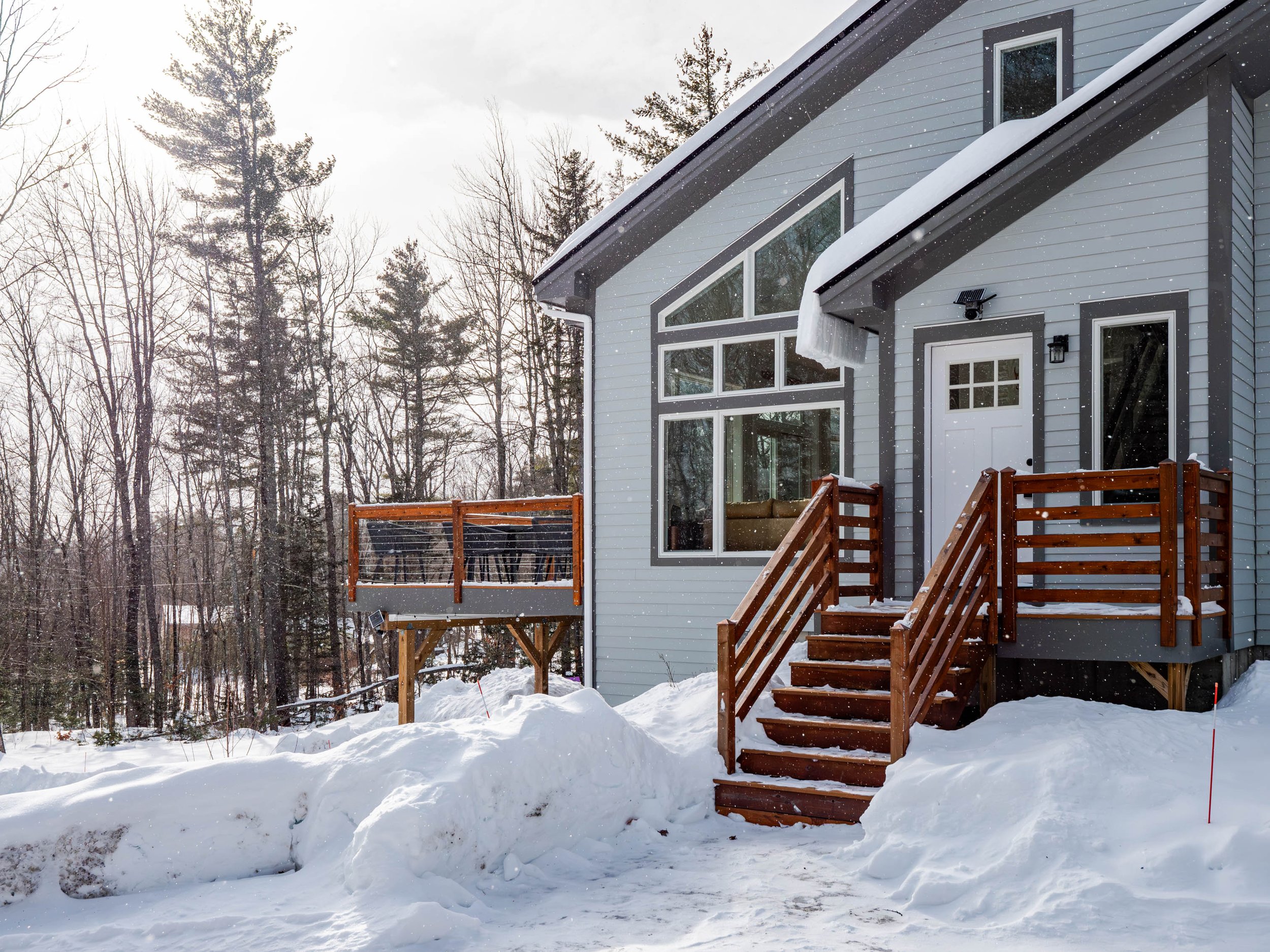 Front of a modern house with snow-covered steps and deck, surrounded by trees, during winter.