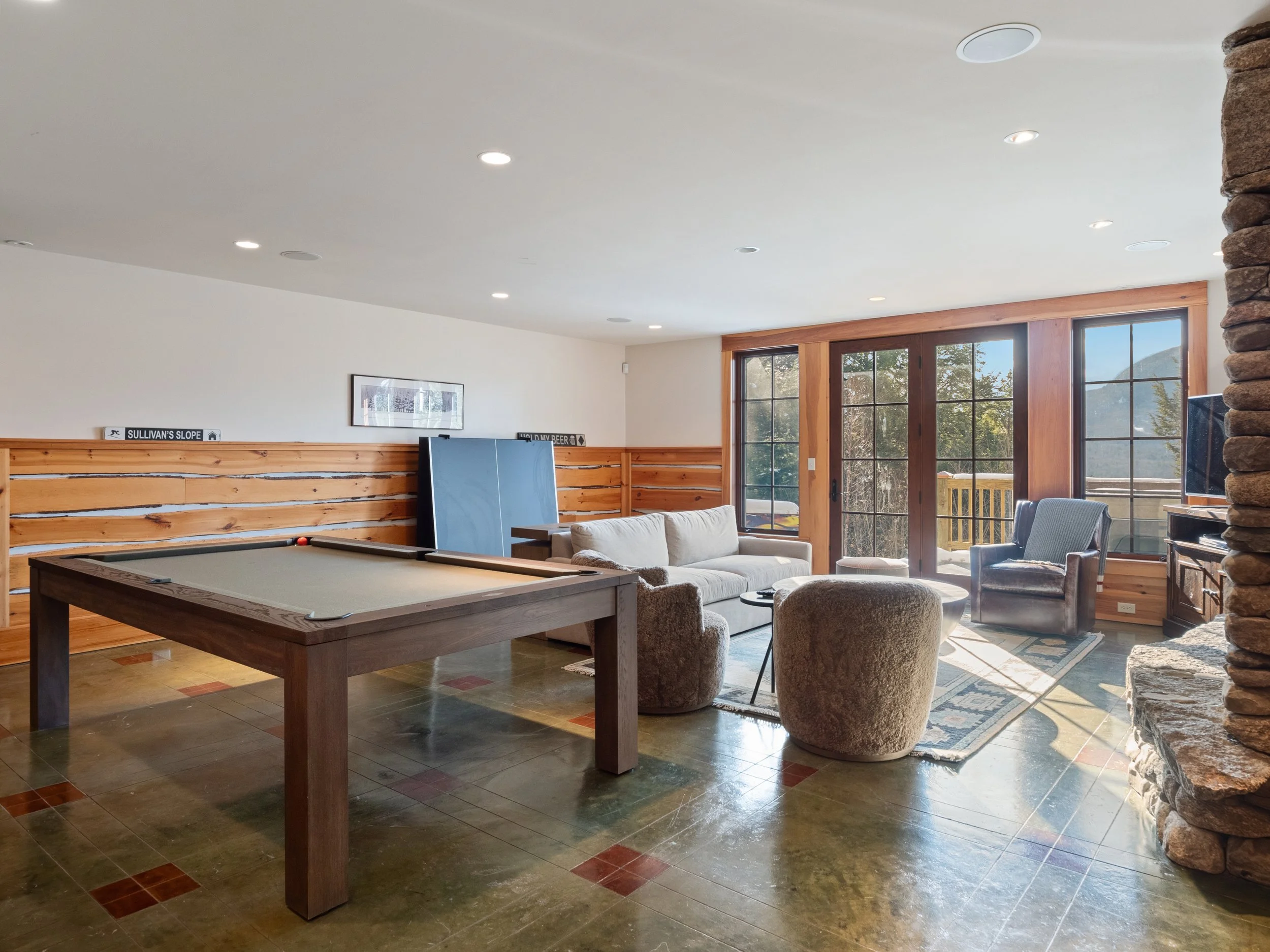 Living room with pool table, sectional sofa, armchair, large windows, and stone fireplace.