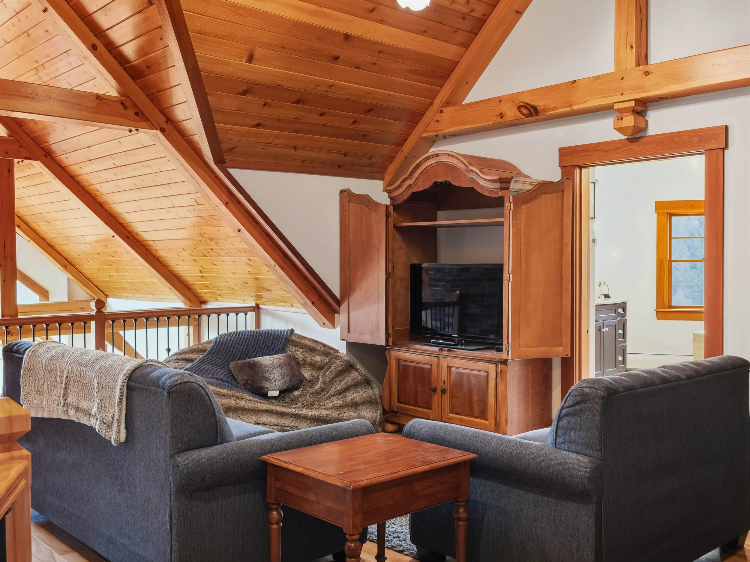 Living room with wooden vaulted ceiling, two gray sofas, a small wooden table, a wooden cabinet with a TV, and an open doorway leading to a bathroom with a window.