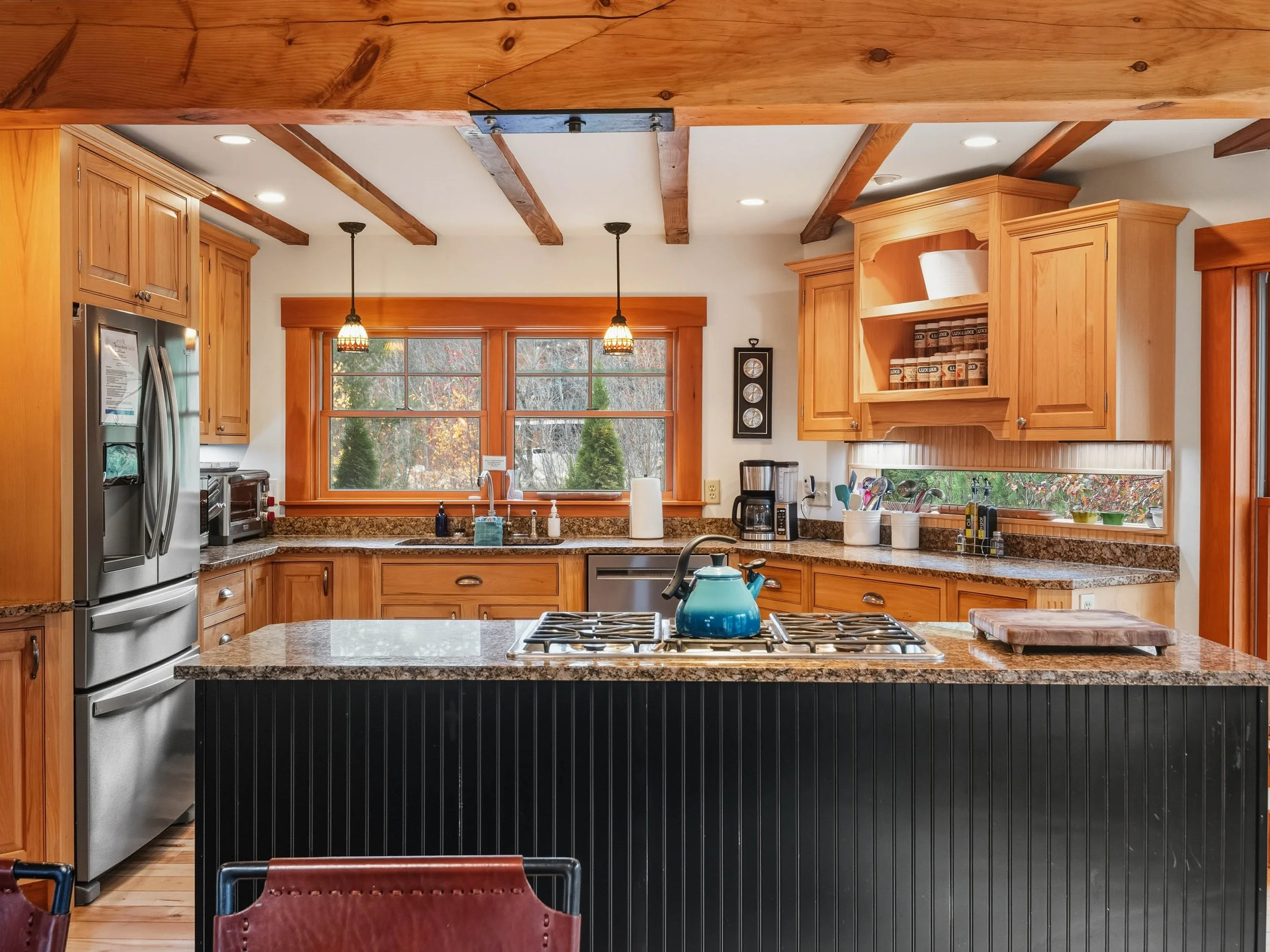 A rustic kitchen with wooden cabinets, granite countertops, and a center island with a blue teapot on it. There are two pendant lights hanging over the window above the sink, which looks out onto a yard with trees. The kitchen also has stainless stee