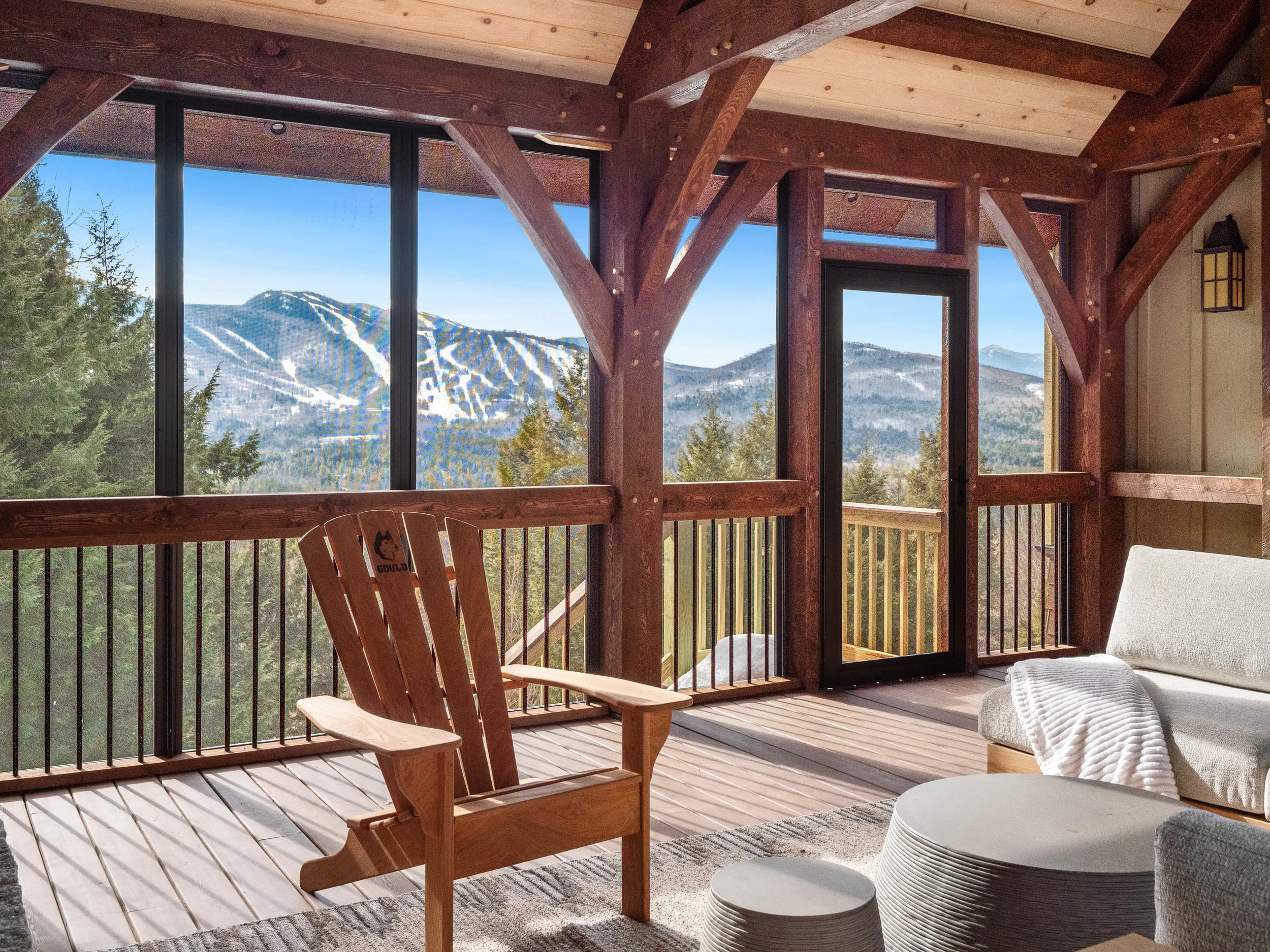 A cozy outdoor balcony overlooking snow-capped mountains and green pine trees, furnished with wooden Adirondack and cushioned seating, and featuring glass doors and wooden beams.