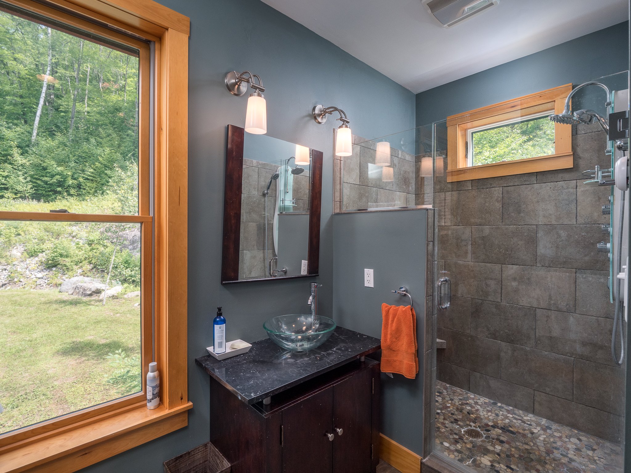Modern bathroom with glass shower, stone tile, glass vessel sink, and window view of green landscape.