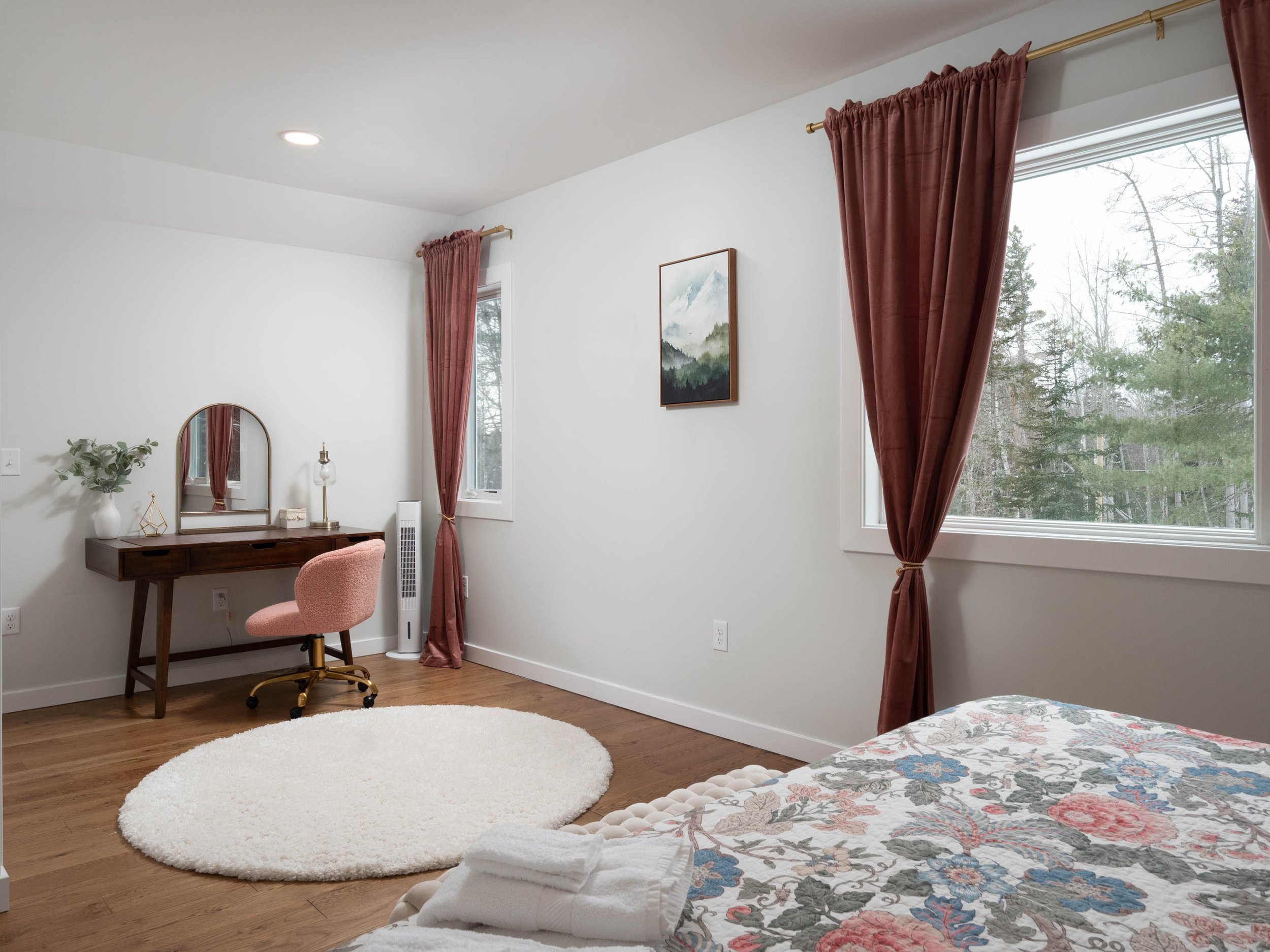 A bedroom with a wooden desk and pink upholstered chair, mirror, night lamp, potted plant, white shag rug, bed with floral quilt, and windows with pink curtains showing trees outside.