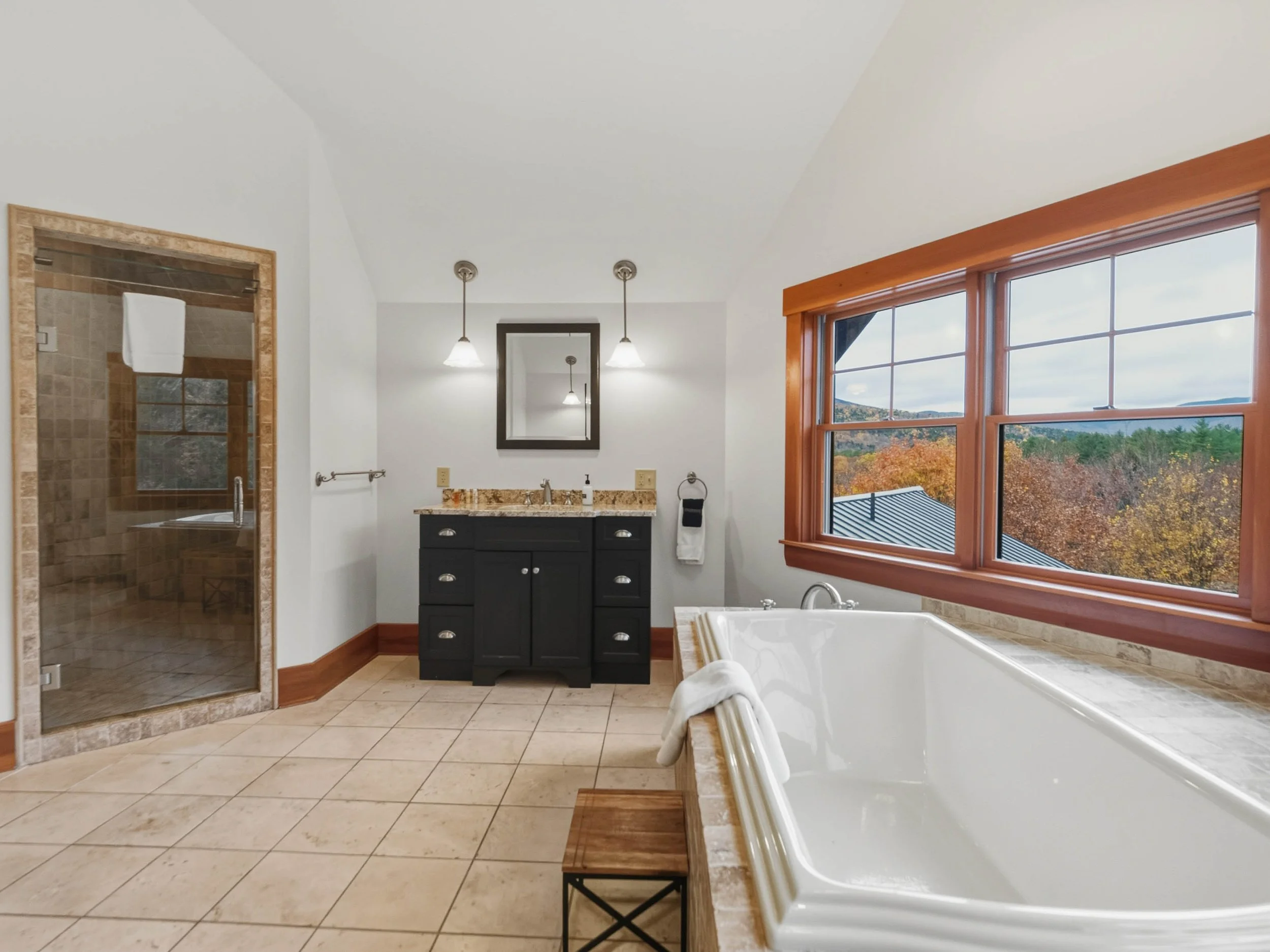 Bathroom with a large window showing an outdoor view, a bathtub, a black vanity with a mirror, a glass-enclosed shower, and tile flooring.