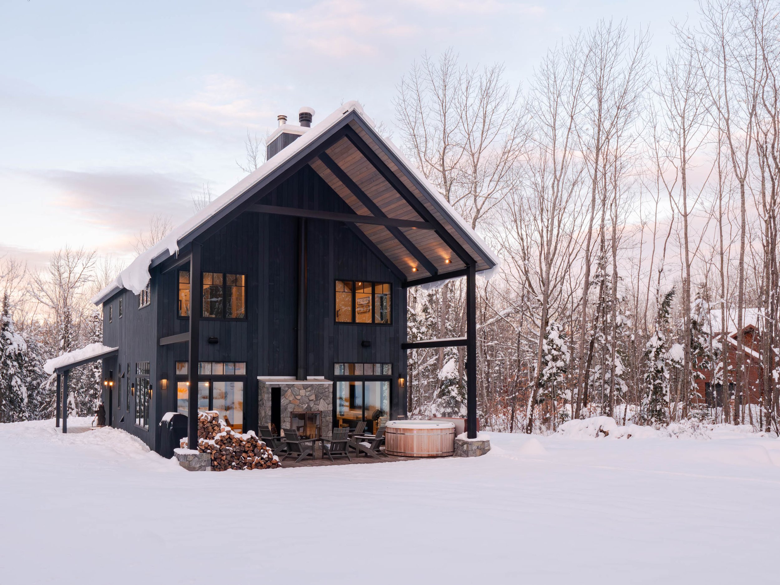 Snow-covered modern cabin in winter forest setting, with large windows, firewood pile, and outdoor fireplace, under a clear sky.