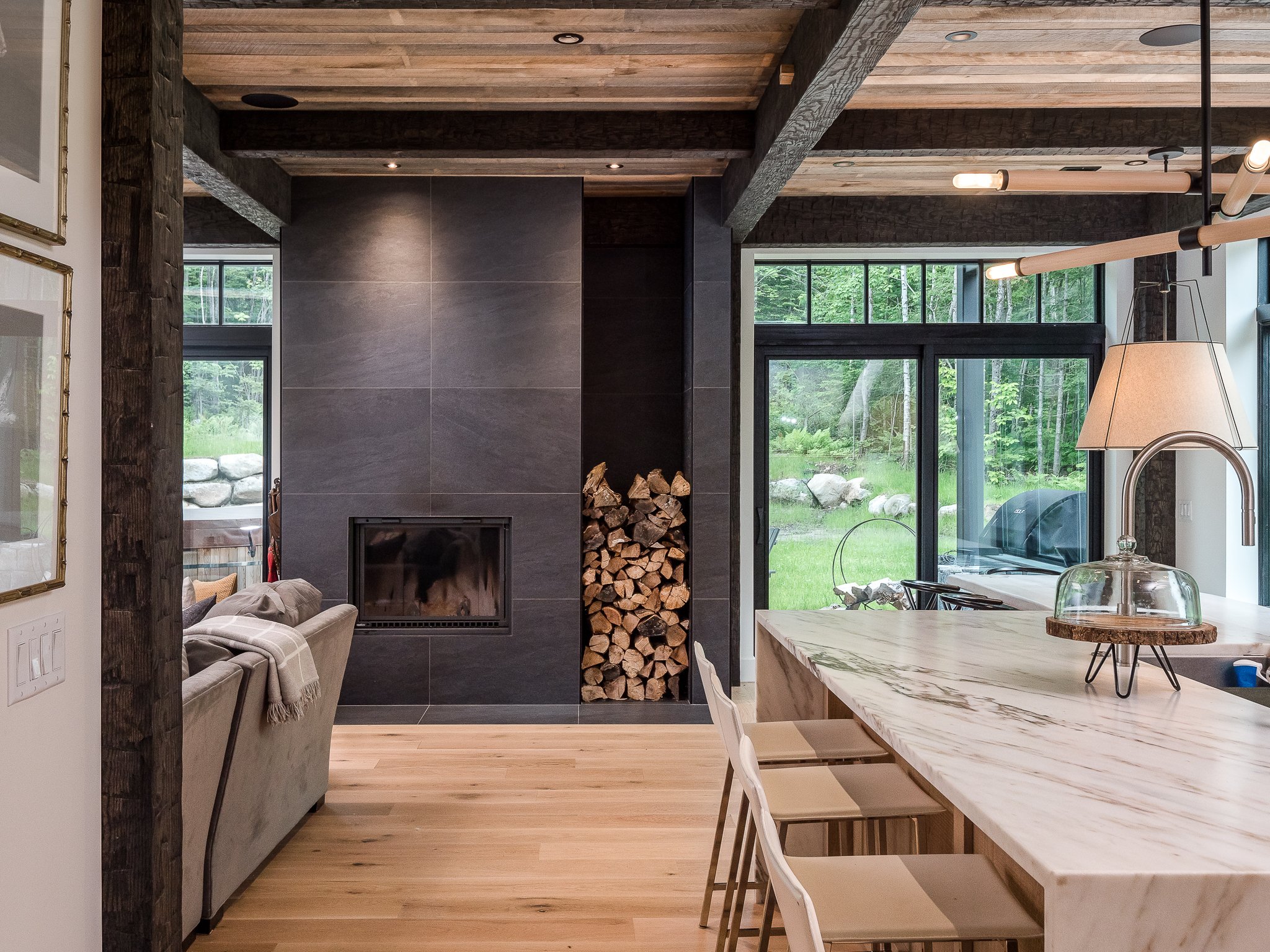 Modern living room with fireplace and large windows, featuring a marble kitchen island, wooden ceiling beams, and stacked firewood.