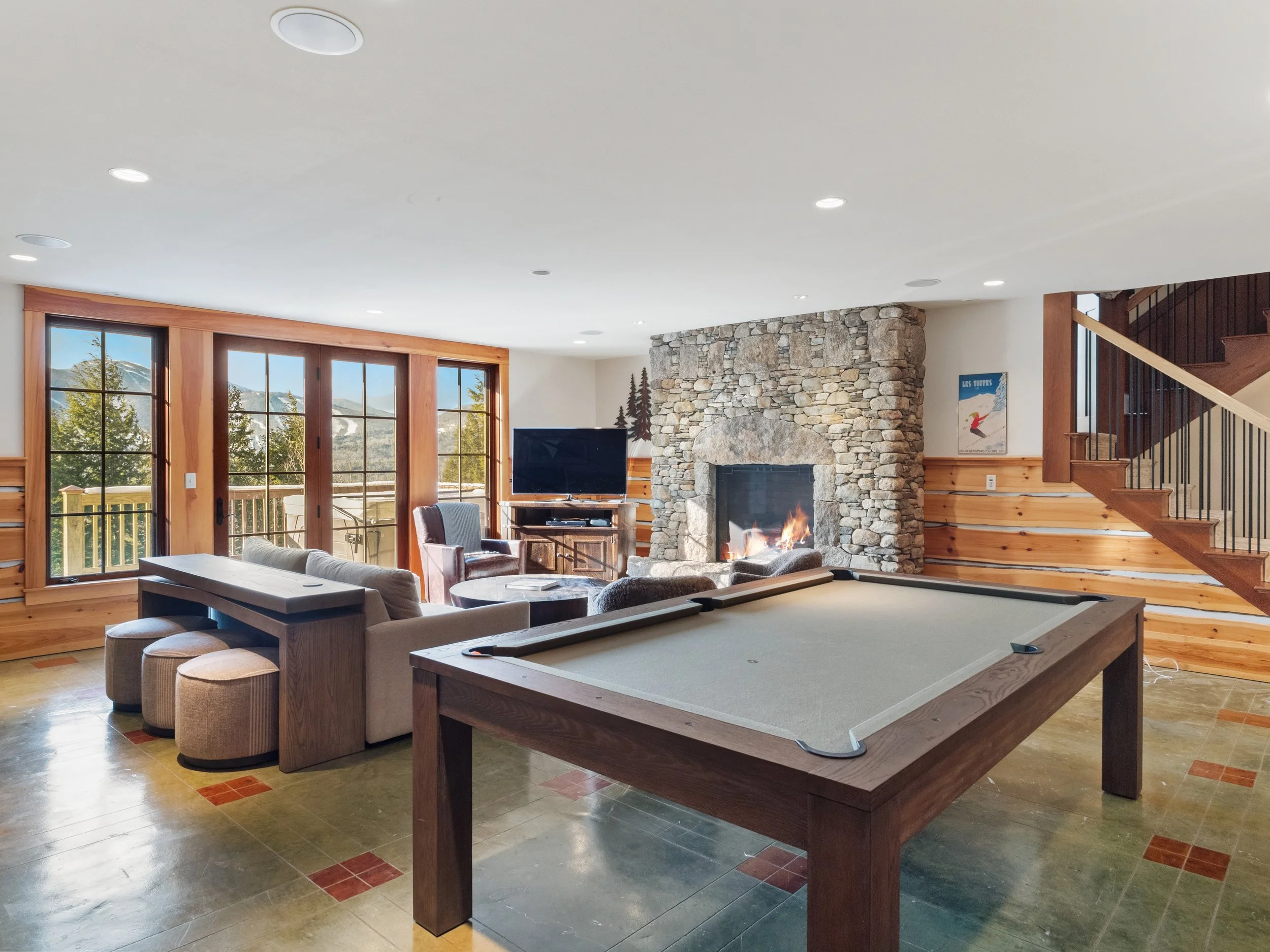 Living room with large windows, a stone fireplace with a fire burning, a pool table, a TV, a couch set, and a staircase.