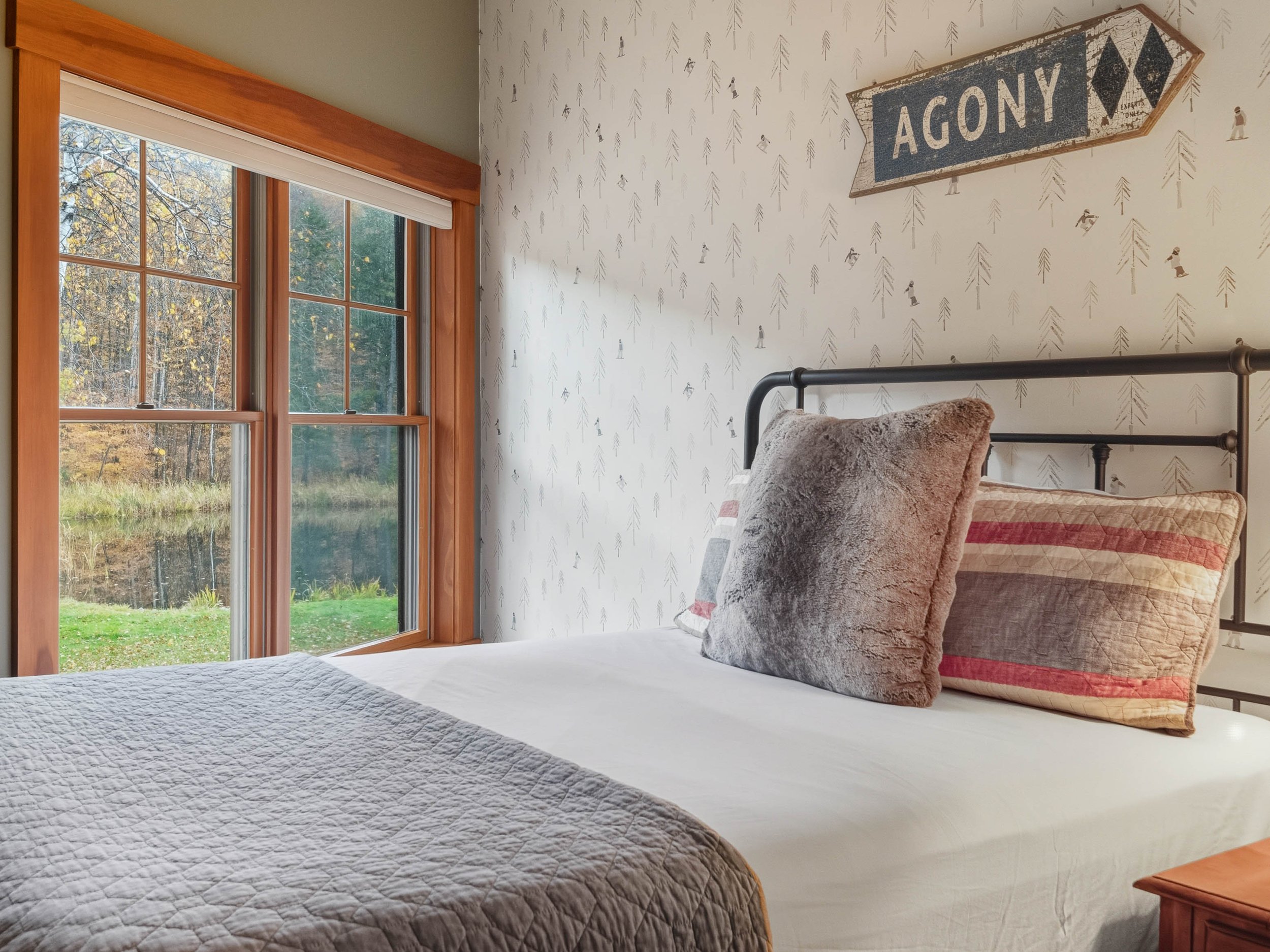 A cozy bedroom with a bed featuring a beige quilt and two pillows, one furry and one with red and beige stripes, near a large window showing trees and a lake outside, and a wooden nightstand beside the bed, with a vintage sign reading 'AGONY' on the 