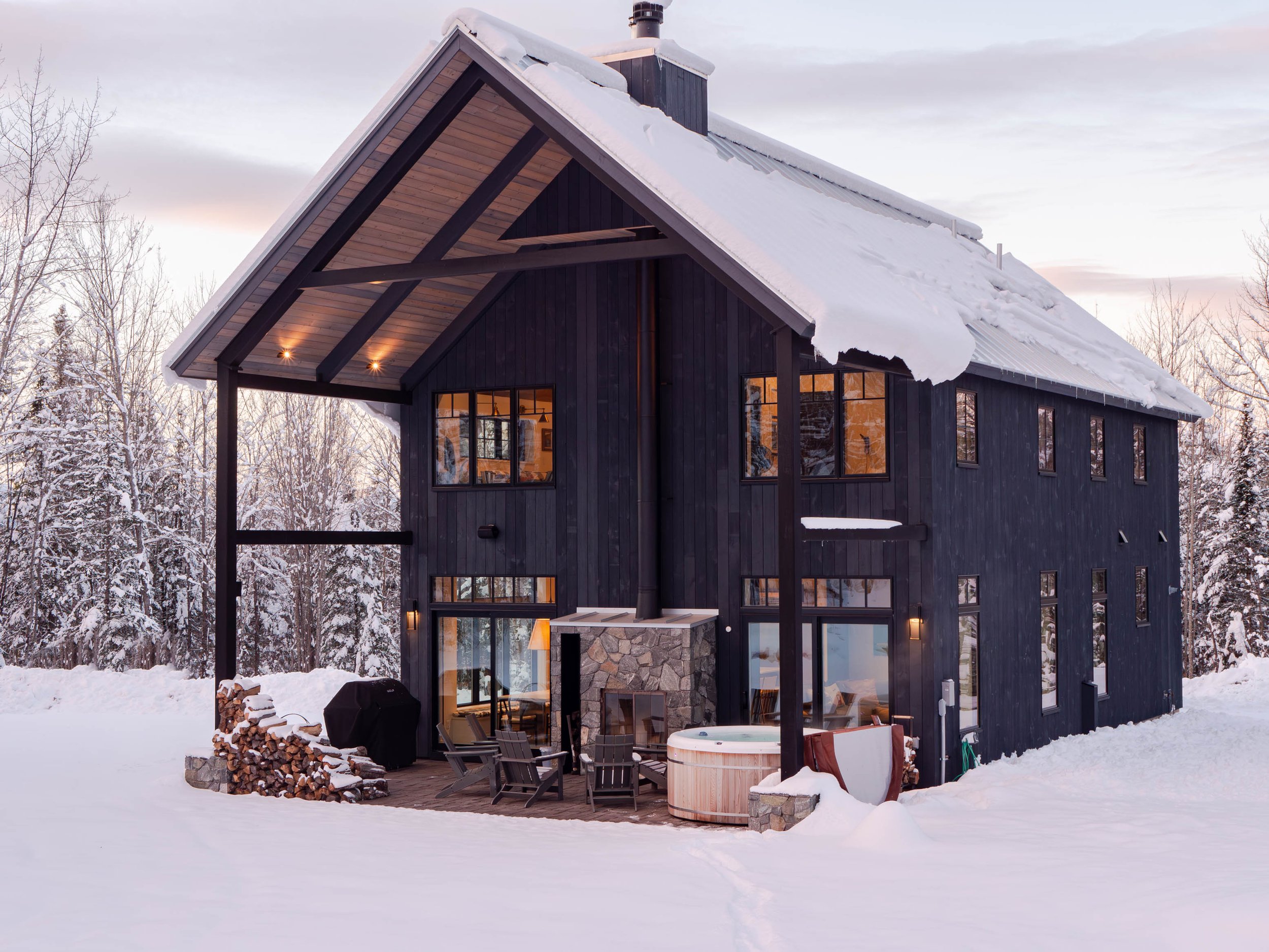 Black modern cabin in snowy landscape with large windows, outdoor firewood stack, and hot tub on patio.
