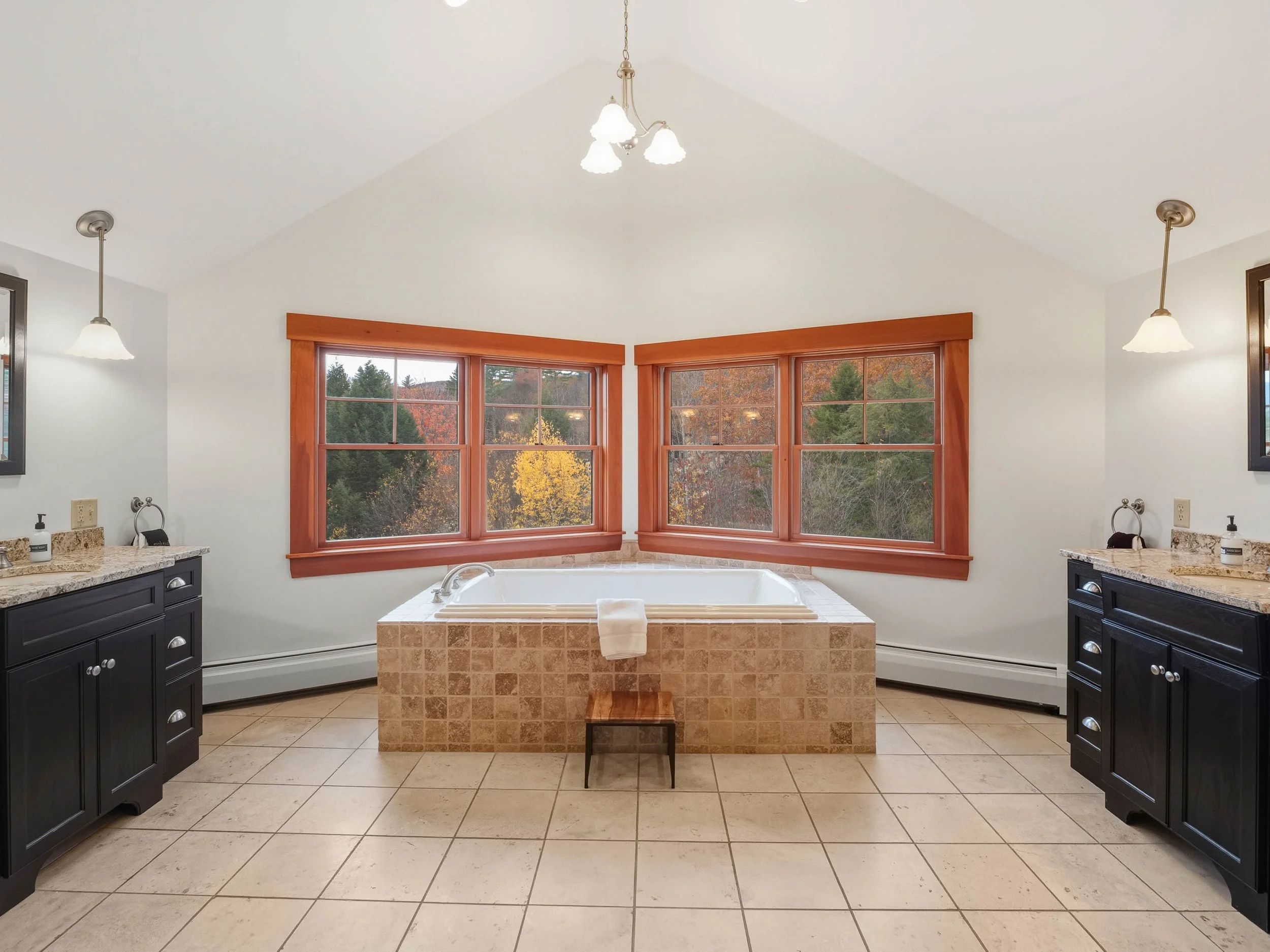 Bathroom with a central bathtub surrounded by windows showing trees with fall foliage, black vanities with granite countertops, bathroom fixtures, and pendant lighting.