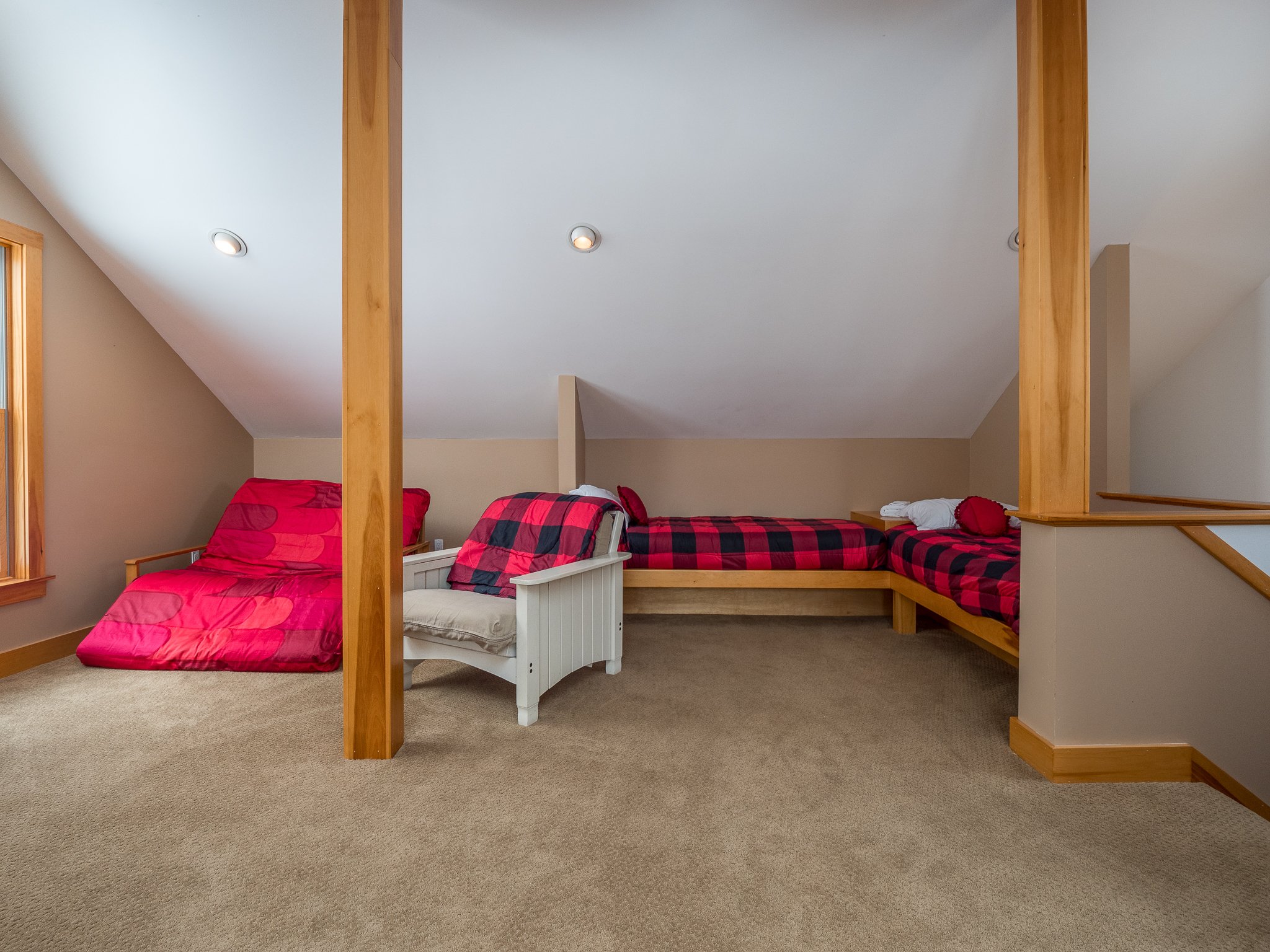 Loft bedroom with two beds featuring red and black checkered bedding, beige carpet, wooden beams, and sloped ceiling.