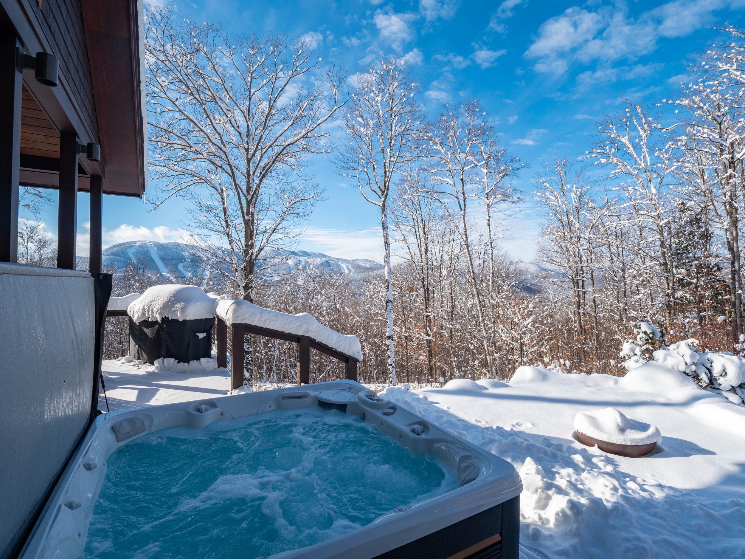 Outdoor hot tub on a snowy deck with snow-covered trees and mountains in the background.