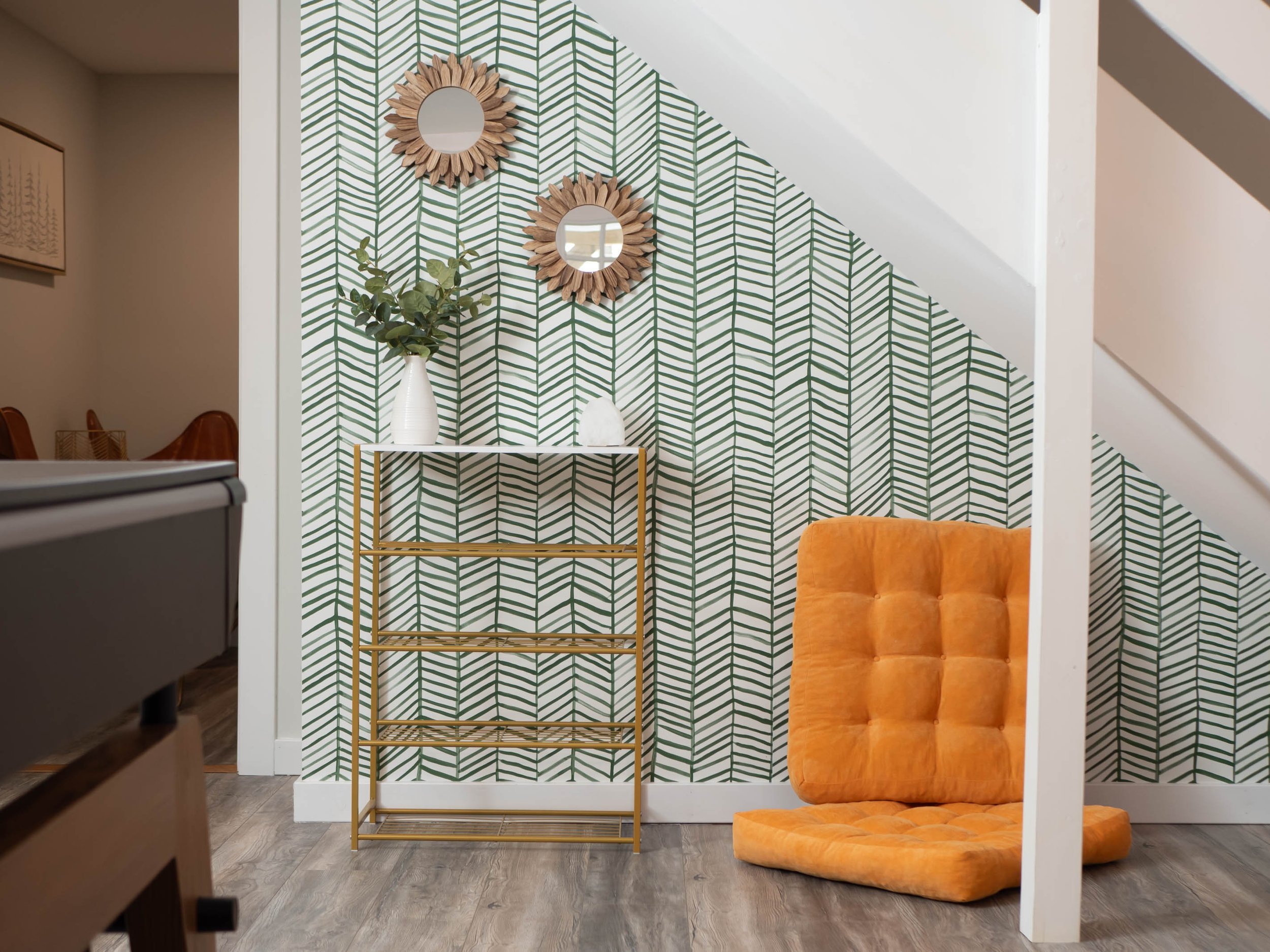 Interior corner with green leaf patterned wallpaper, two round wooden mirrors, an orange floor cushion, a gold metal shelf, a white vase with green leaves, and a white decorative object.