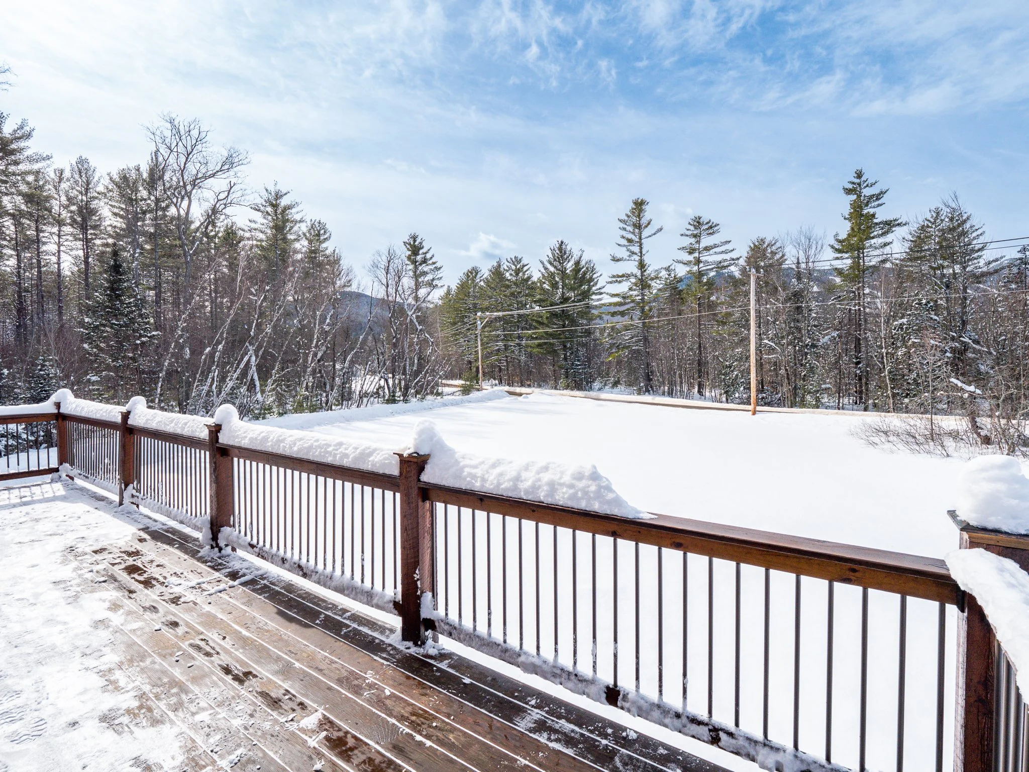 Snow-covered deck with wooden railing overlooking a snowy landscape and trees.