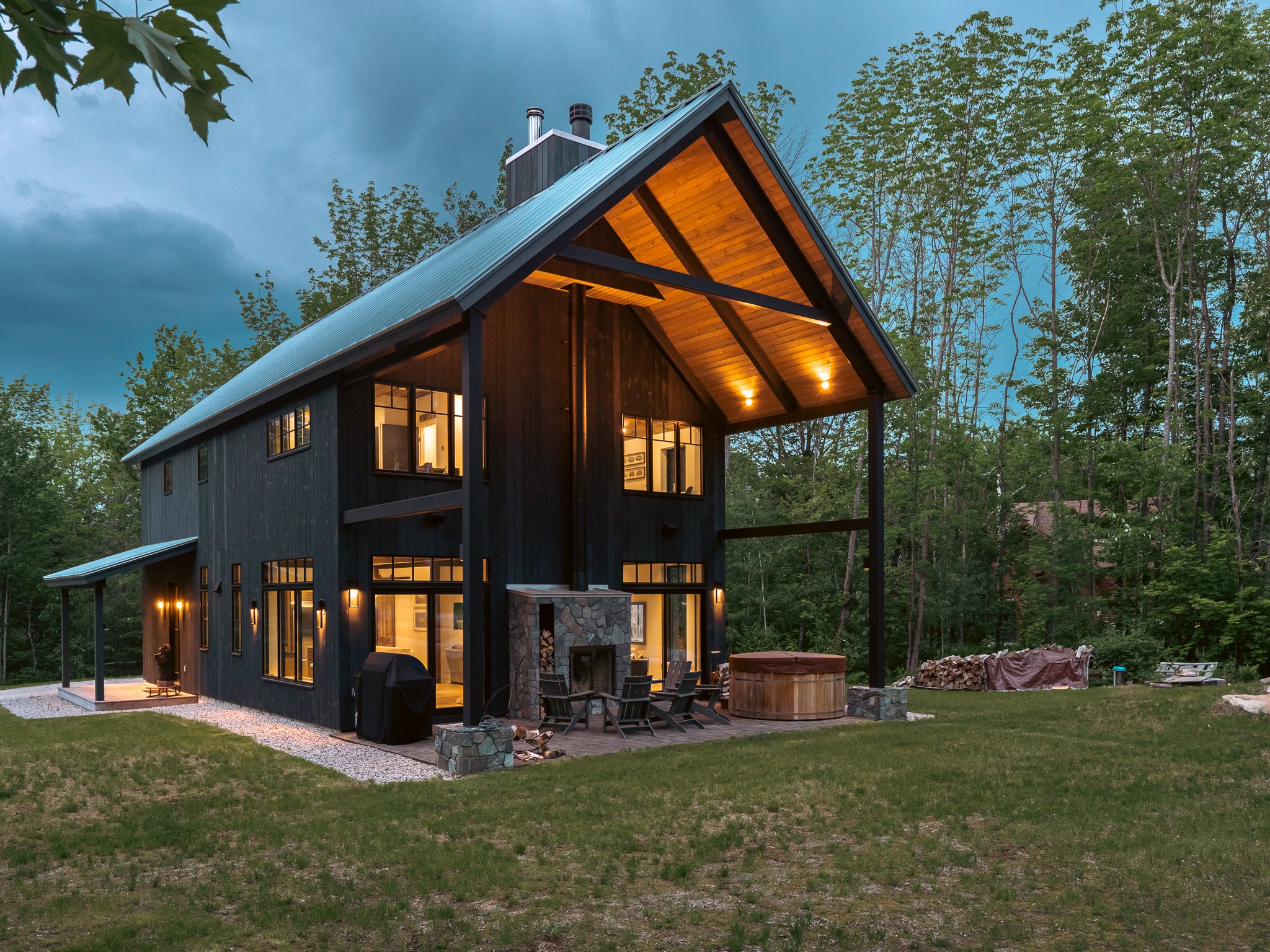 Modern wooden house with large windows and patio, surrounded by trees and lawn, during evening.