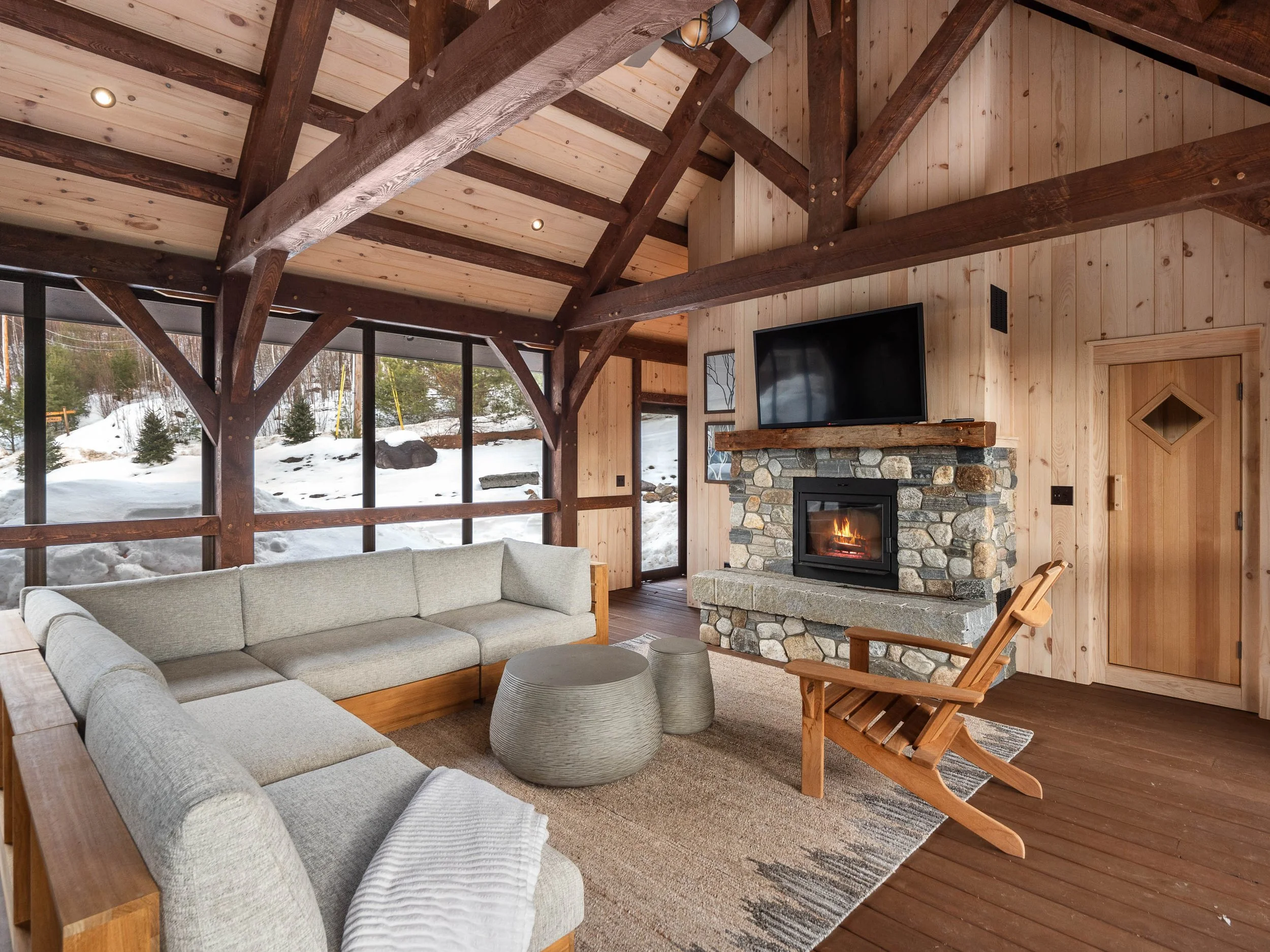 Cozy living room with beige couches, a stone fireplace with a fire, a wooden chair, a TV above the fireplace, and windows showing snow outside.