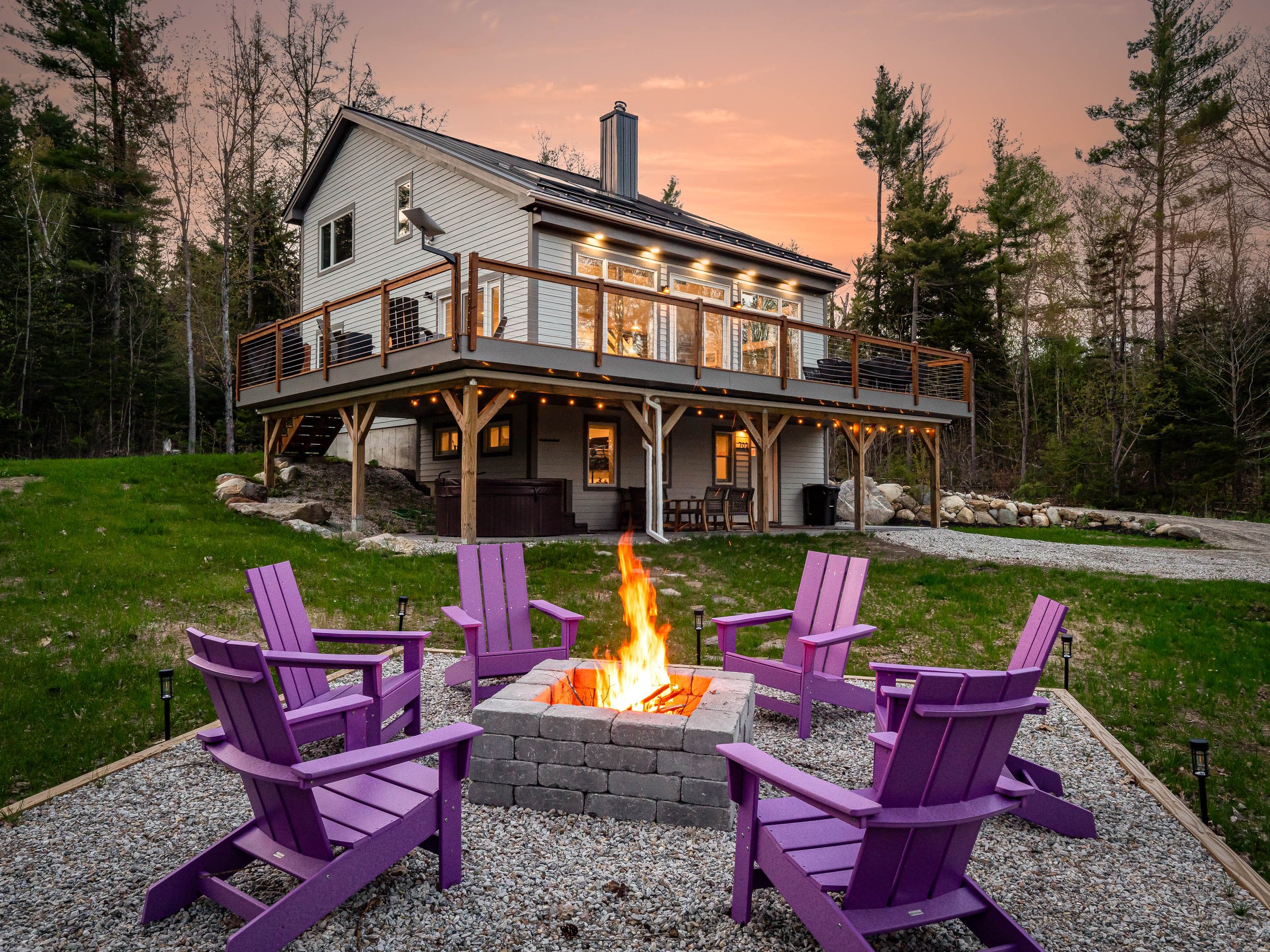 A house with a large outdoor deck and a fire pit surrounded by six purple Adirondack chairs in a grassy yard at sunset.
